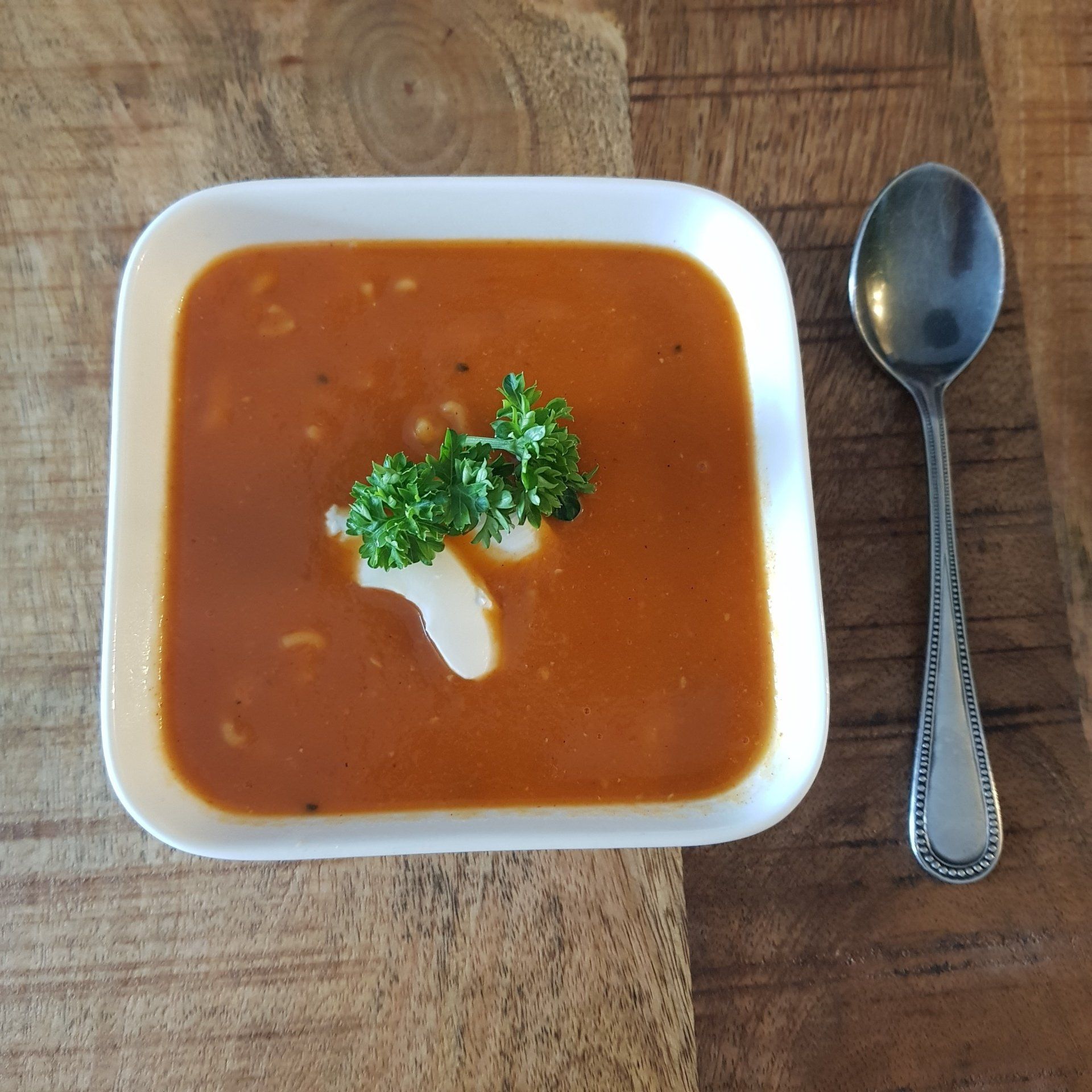 Een kom tomatensoep, gegarneerd met room en peterselie, naast een lepel op een houten tafel.
