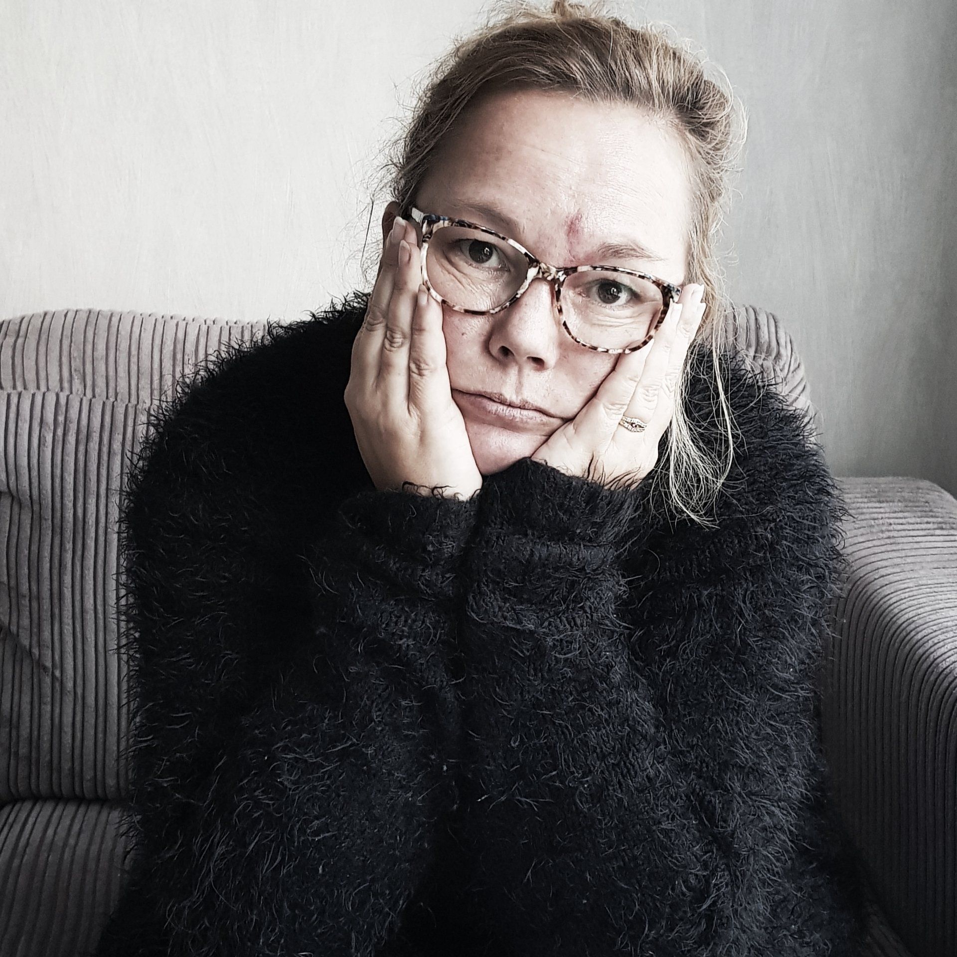 Een vrouw met een bril, haar gezicht in haar handen rustend, er verdrietig uitziend, gekleed in een zwarte trui, zittend op de bank.