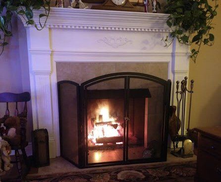 Fireplace with burning logs, black screen, white mantel with greenery, dark chair to the left, tools to the right.