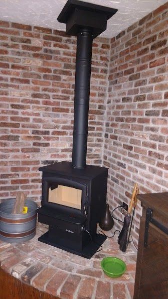 Black wood-burning stove with vertical chimney in a brick-walled corner. Metal bucket and small bowls on brick hearth.