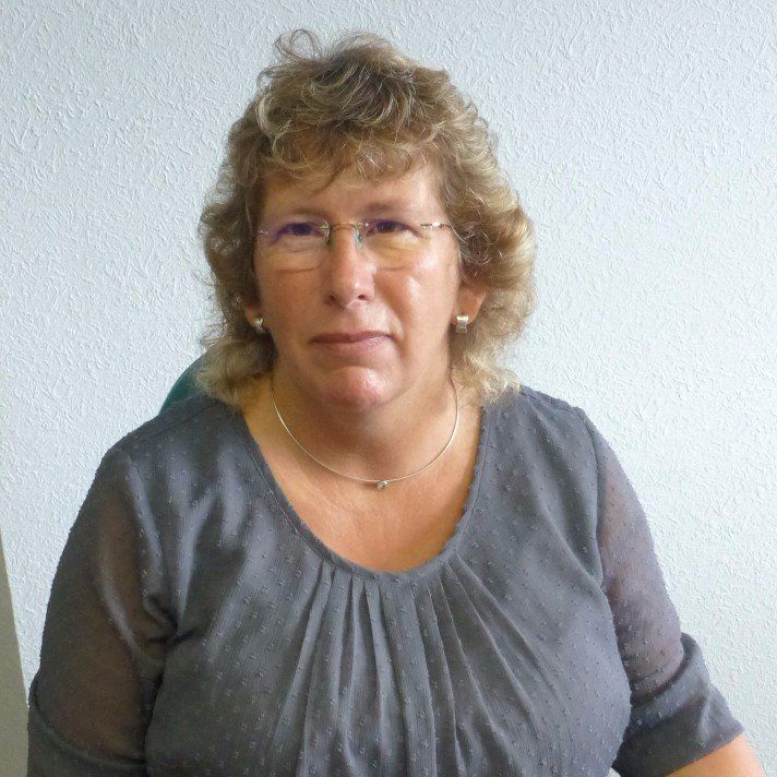 Eine Frau mit Brille und grauem Hemd steht vor einer weißen Wand