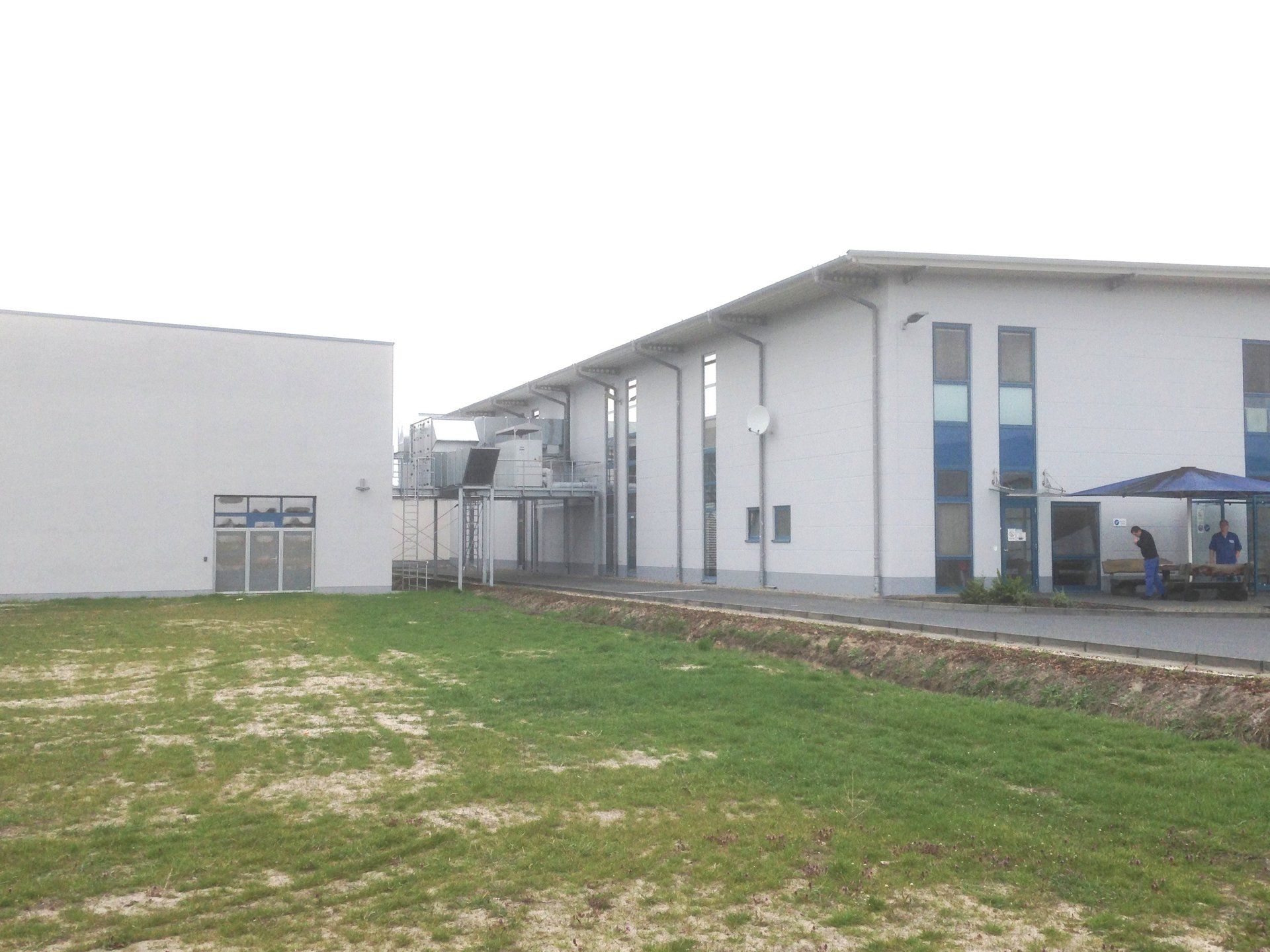 A large white building with blue windows is surrounded by a lush green field.