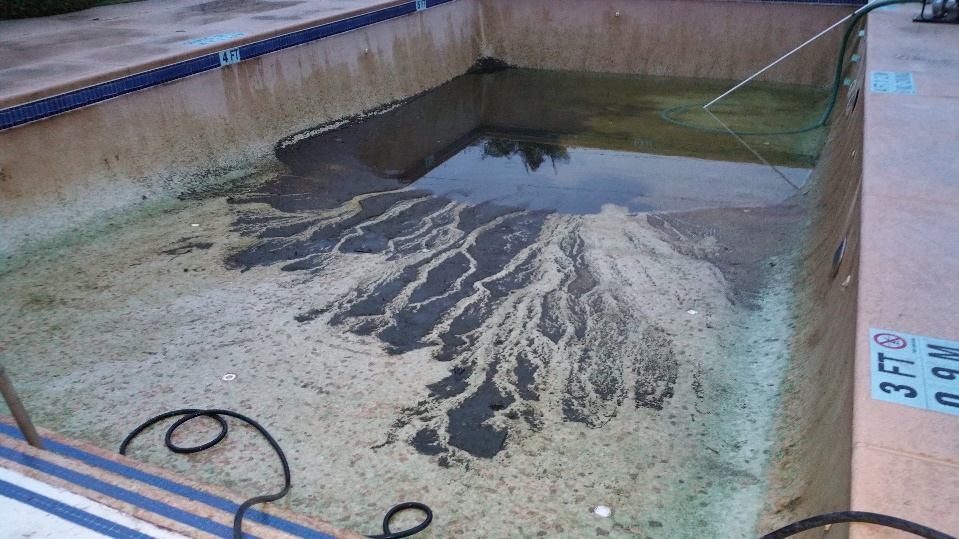 Empty concrete pool with dark water in one corner and a wet, debris-streaked floor.