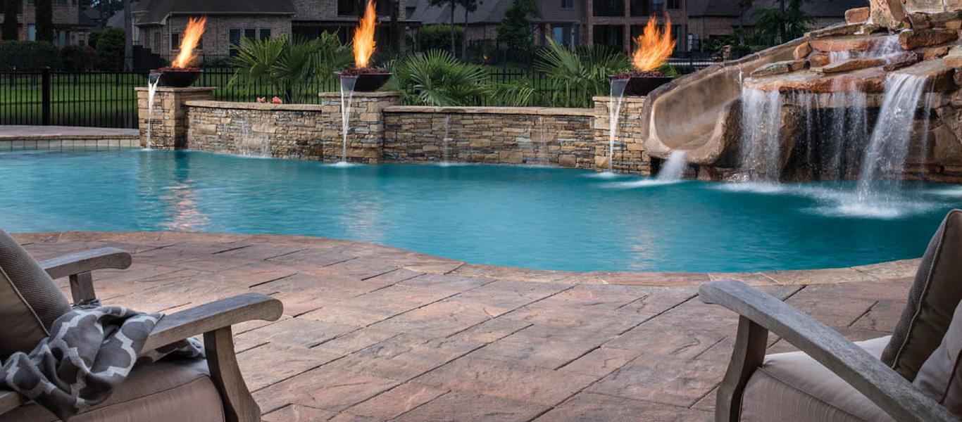 Poolside lounge chairs beside a turquoise pool with a stone waterfall and flames in the background