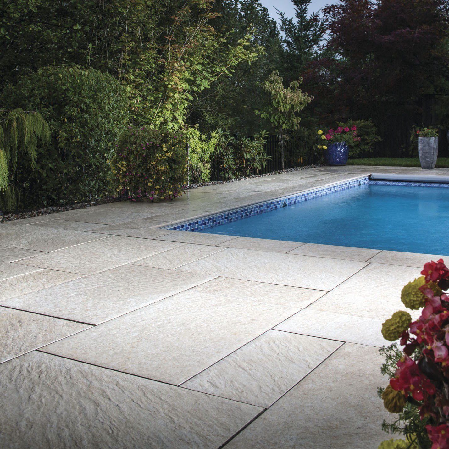 Backyard pool beside a light stone patio, bordered by dense green shrubs and red flowers