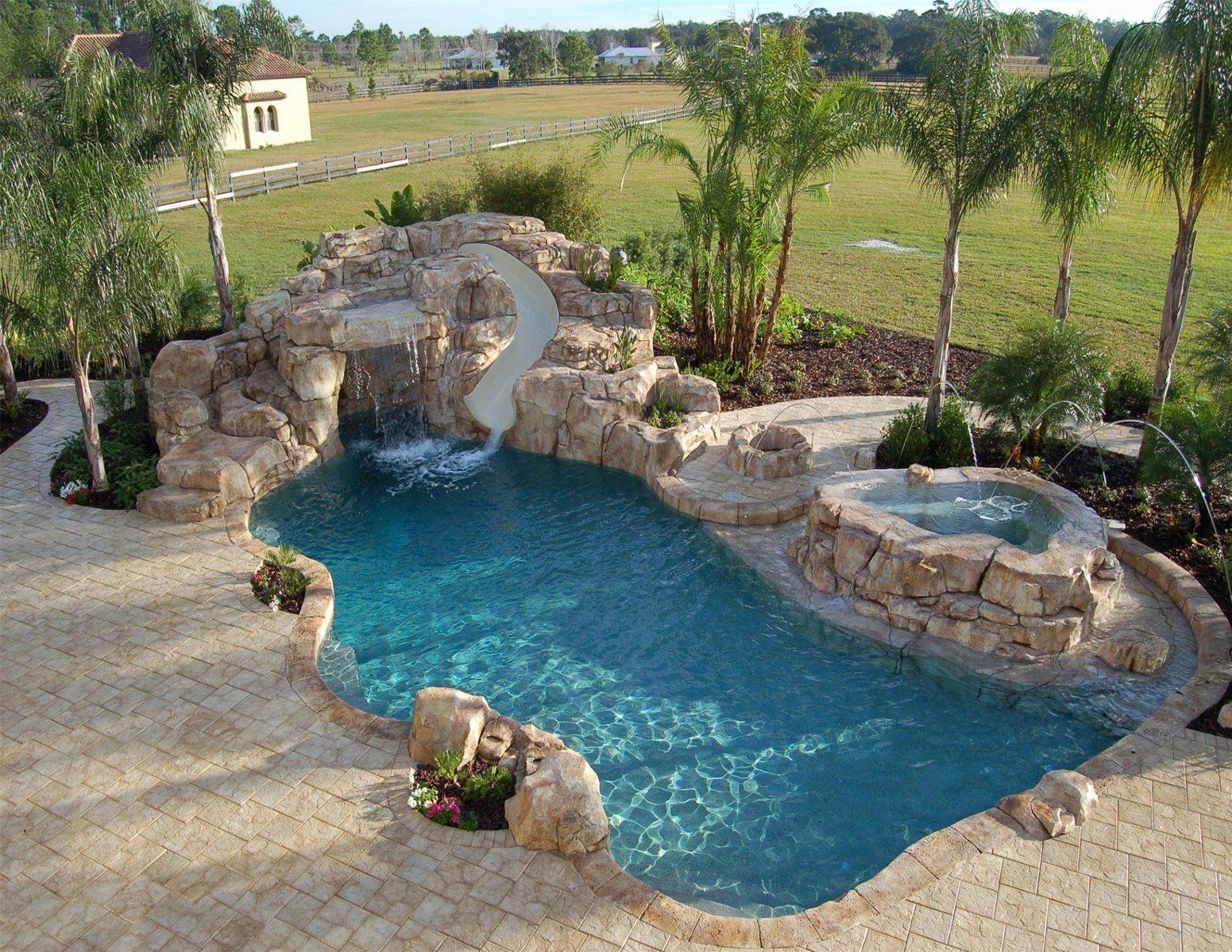 Backyard pool with rock waterfall, blue water, and stone patio surrounded by palm trees
