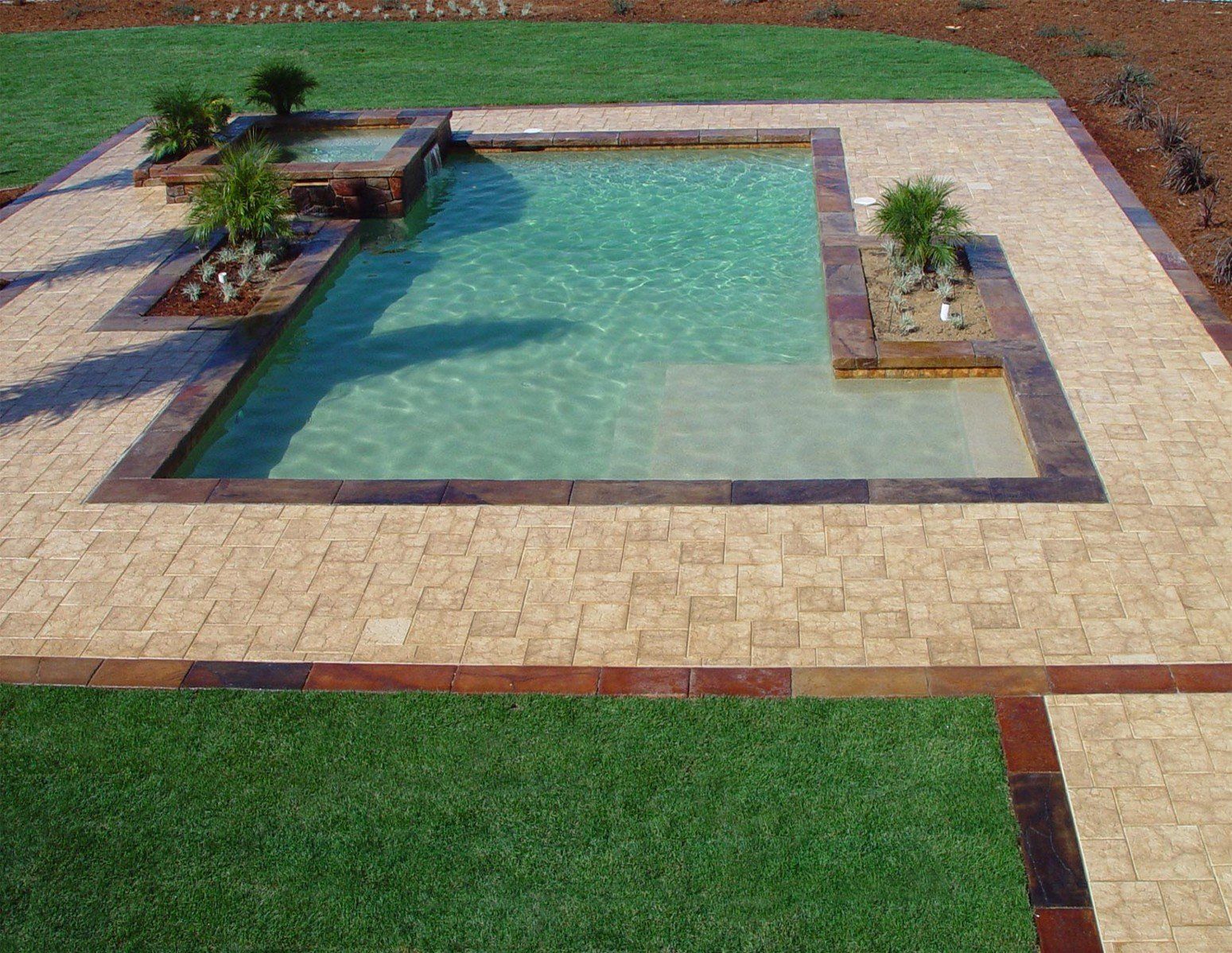 Small backyard swimming pool with tan pavers, green lawn, and potted plants around the deck.