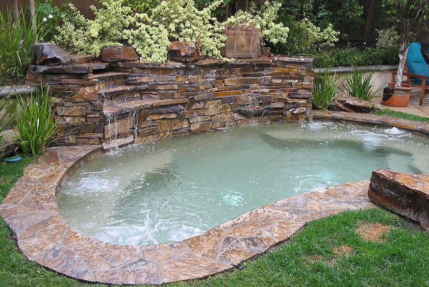 Stone-lined backyard hot tub with bubbling water and a stacked rock waterfall, surrounded by greenery.