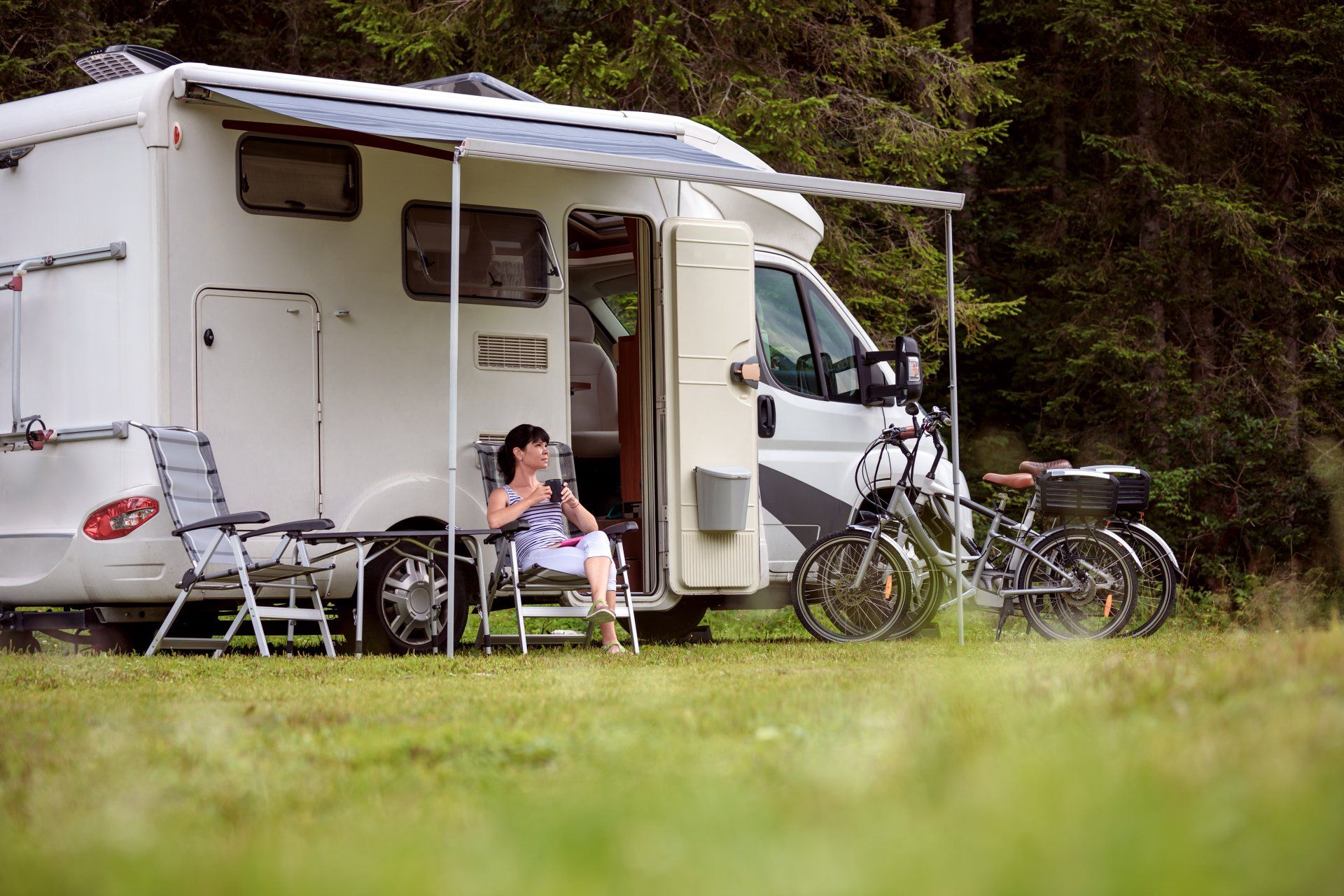 Person relaxing outside a white RV with awning, two bicycles, and camping chairs in a grassy area with trees.