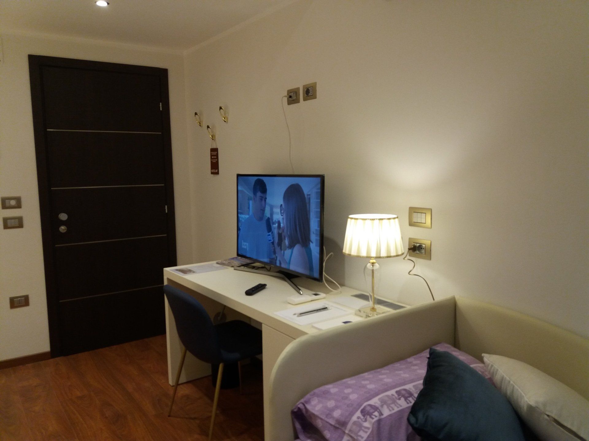 Piccola stanza con scrivania, TV e lampada vicino al letto. Porta marrone scuro sulla sinistra, pareti bianche e pavimento in legno.