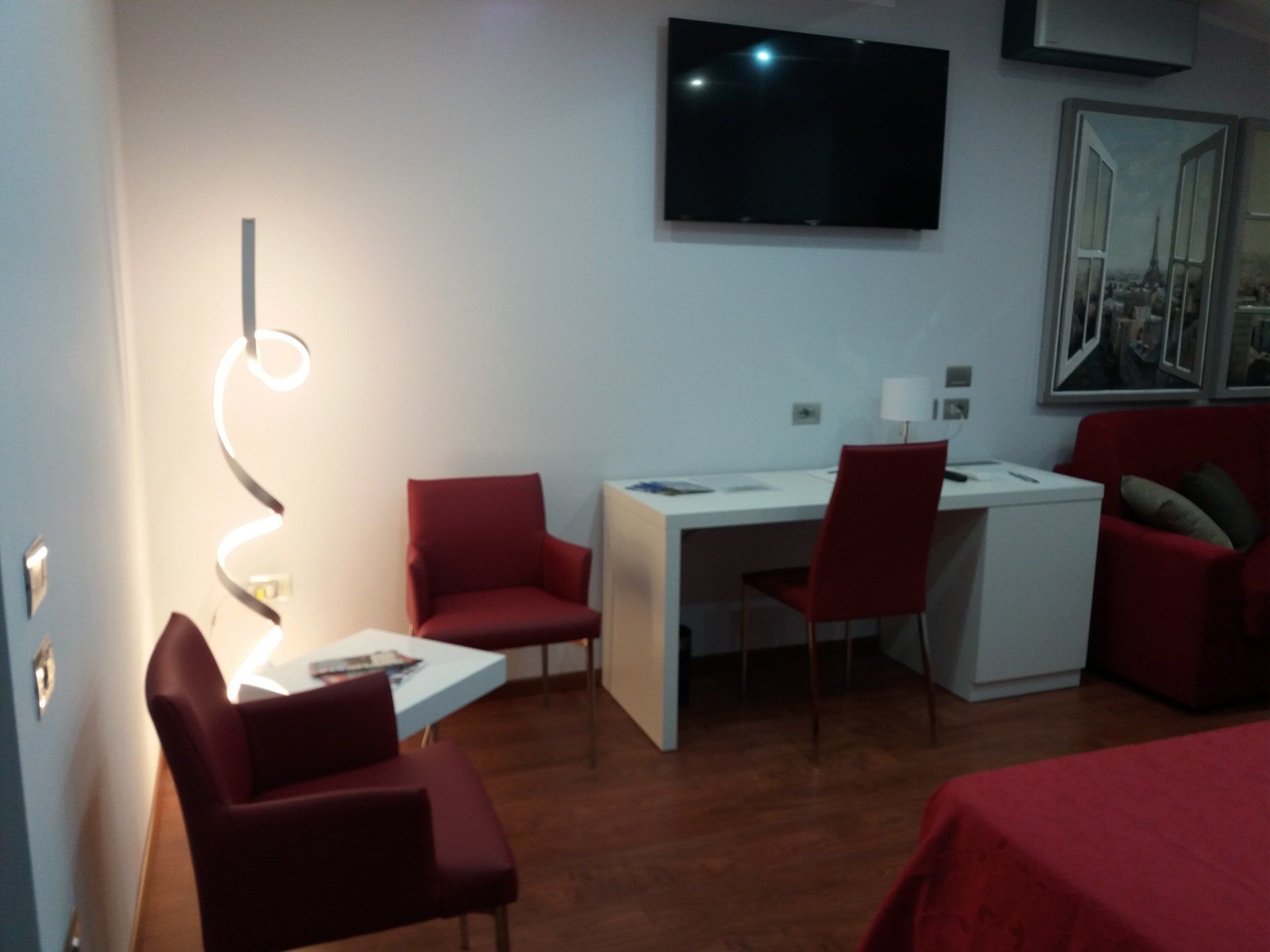 Una moderna camera d'albergo con dettagli rossi: sedie, sedia da scrivania e letto. Una scrivania bianca, TV e lampada decorativa.