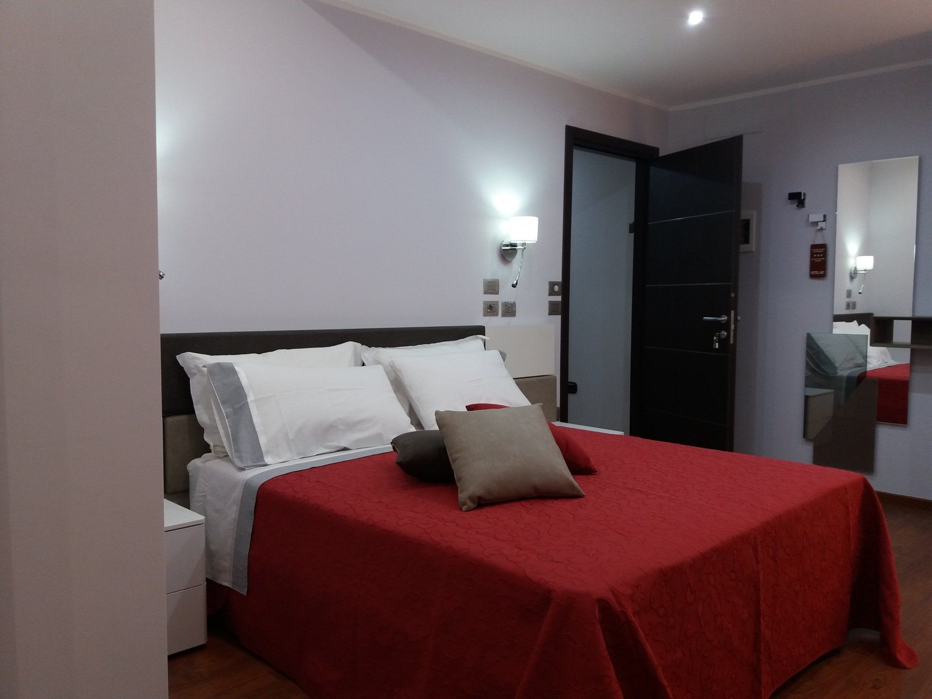 Camera da letto con letto coperto da una coperta rossa, cuscini bianchi e testiera marrone scuro. Porta nera e specchio.