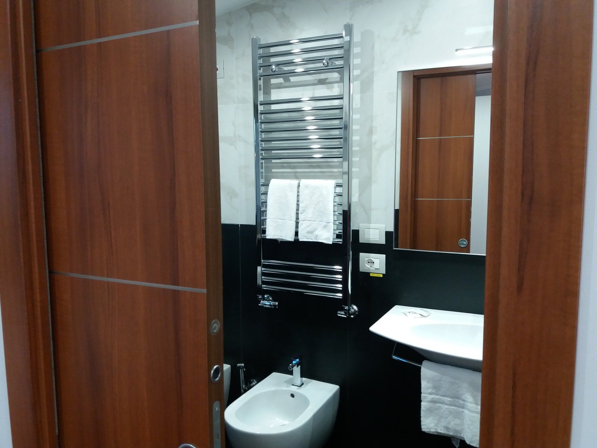 Bagno con porta marrone scuro, portasciugamani cromato, lavandino bianco, bidet e specchio.