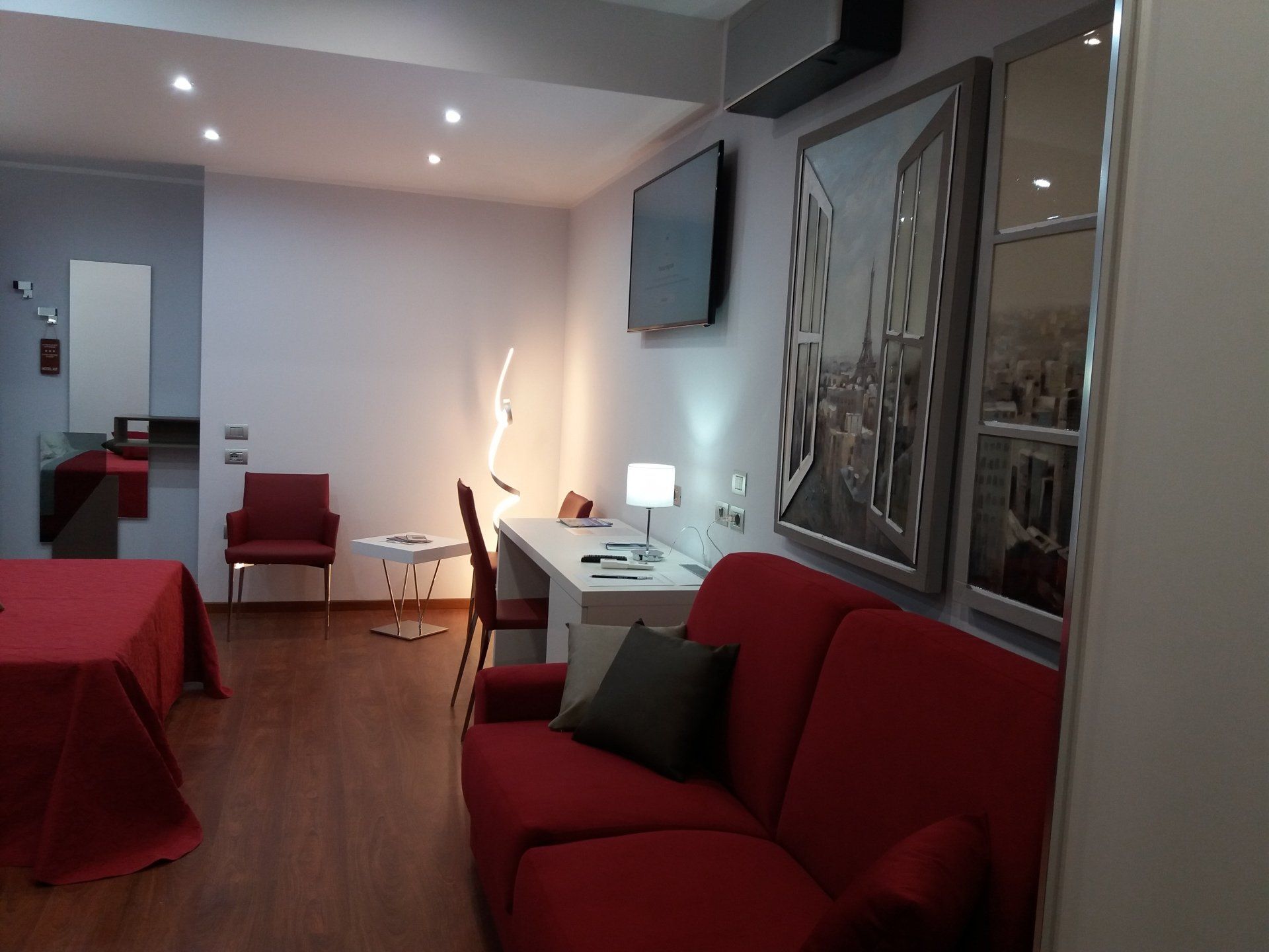 Camera d'albergo con divano rosso, letto, scrivania e quadri raffiguranti paesaggi urbani. Pavimento in legno scuro e pareti grigie.