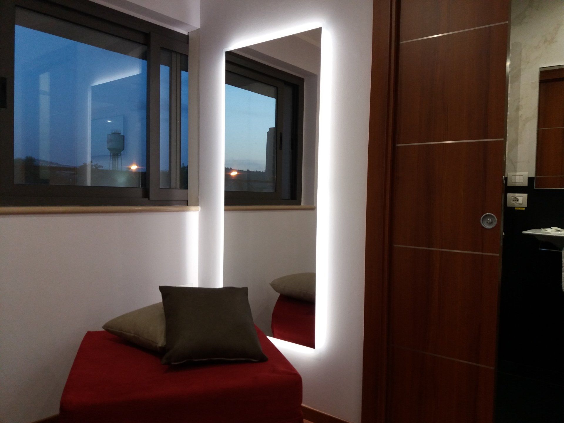 Specchio con illuminazione a LED su una parete accanto a una finestra e una porta; davanti allo specchio c'è un pouf rosso con cuscini.