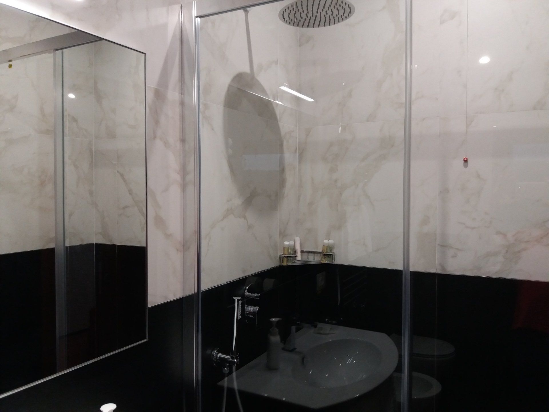 Bagno con doccia in vetro, lavandino bianco, piastrelle in marmo e grande specchio. Combinazione di colori bianco e nero.