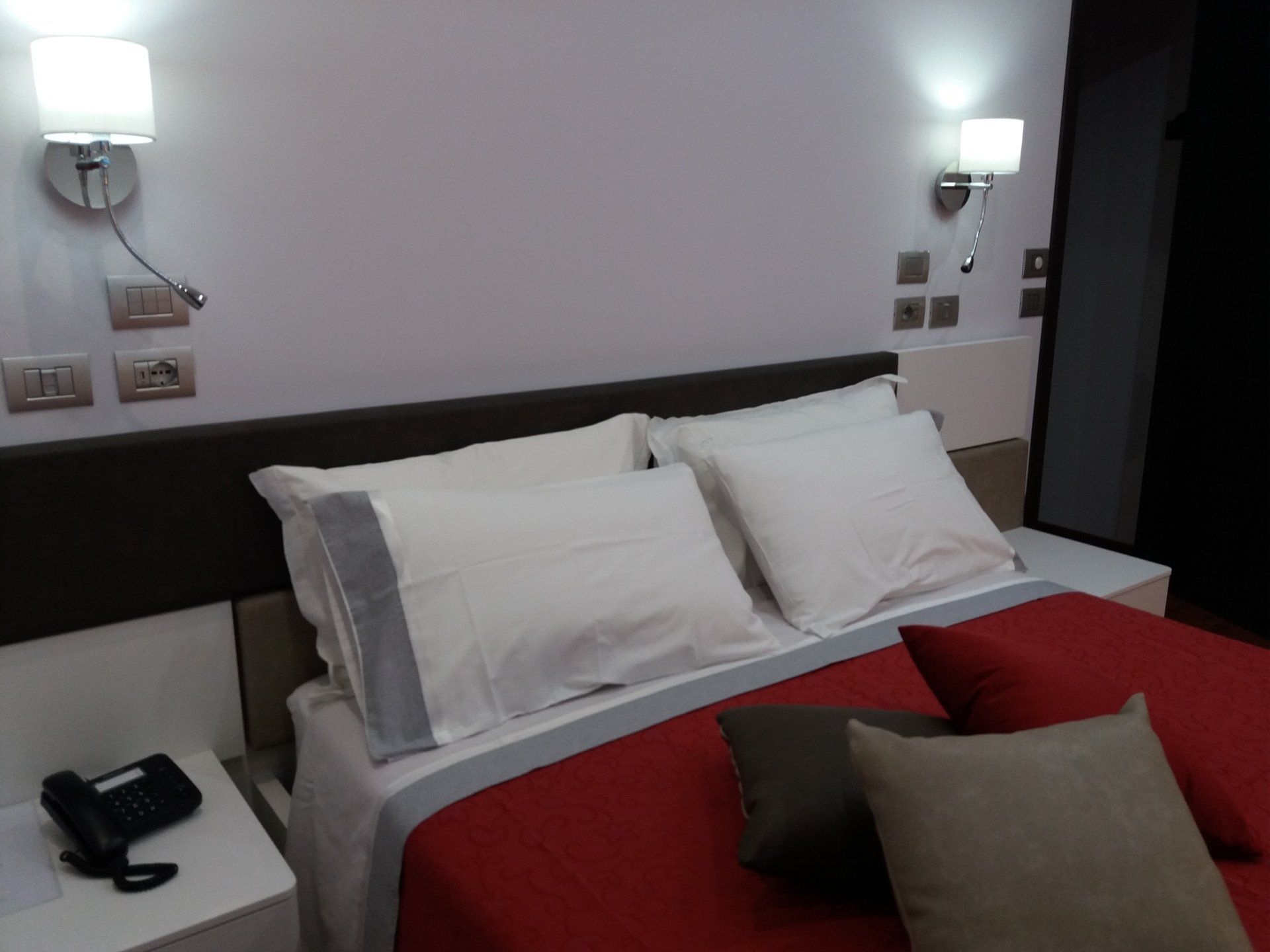 Camera da letto con copriletto rosso, cuscini bianchi e applique.