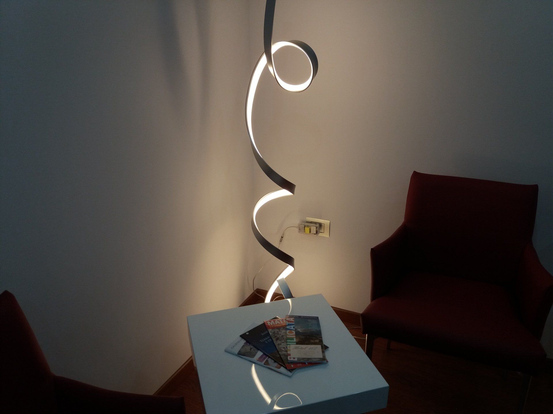 Una moderna lampada da terra con design a spirale illumina un tavolo bianco e due sedie rosse.