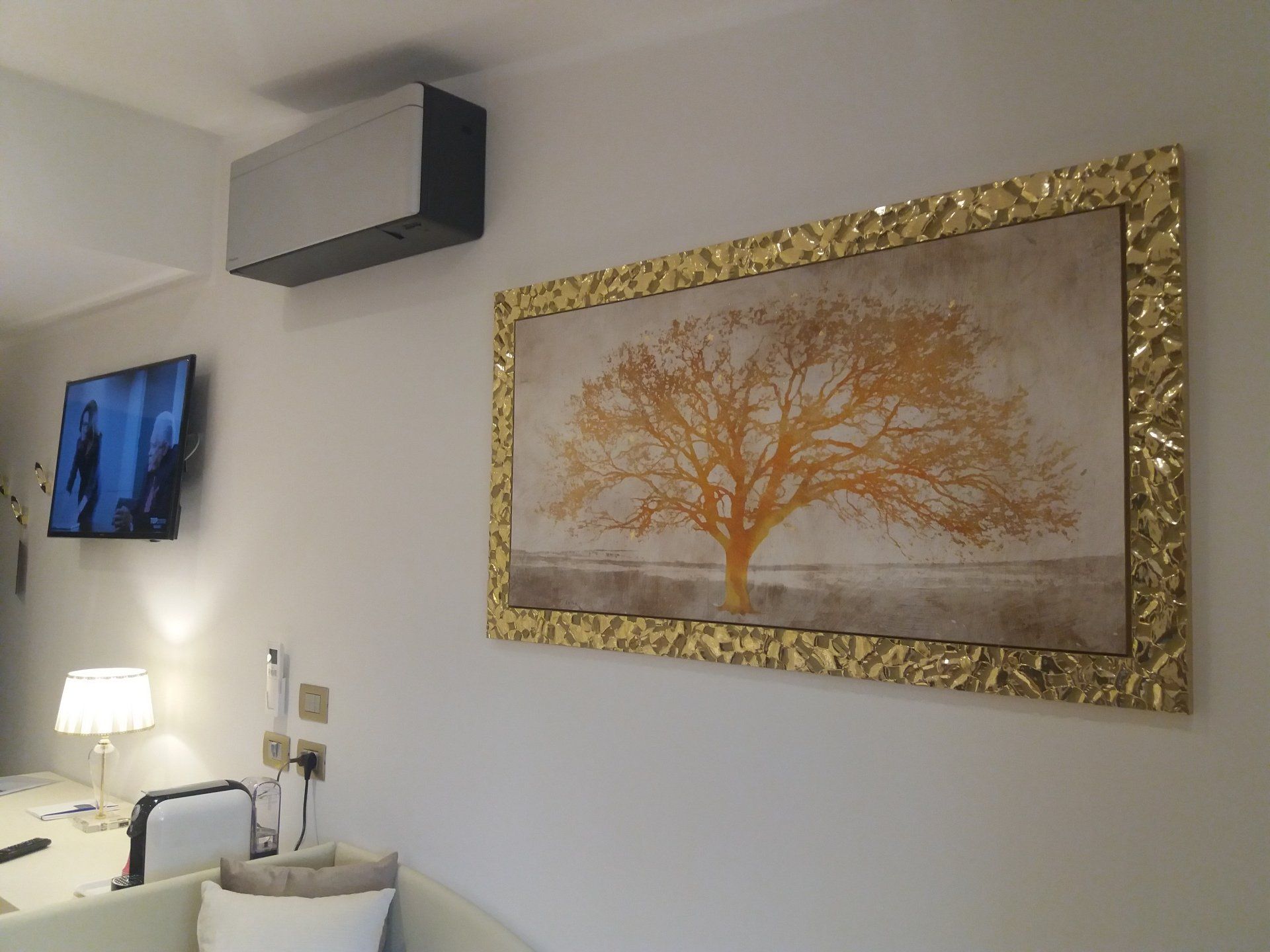 Stanza con opere d'arte e TV a parete, un condizionatore sopra le opere d'arte e un tavolino con una lampada.