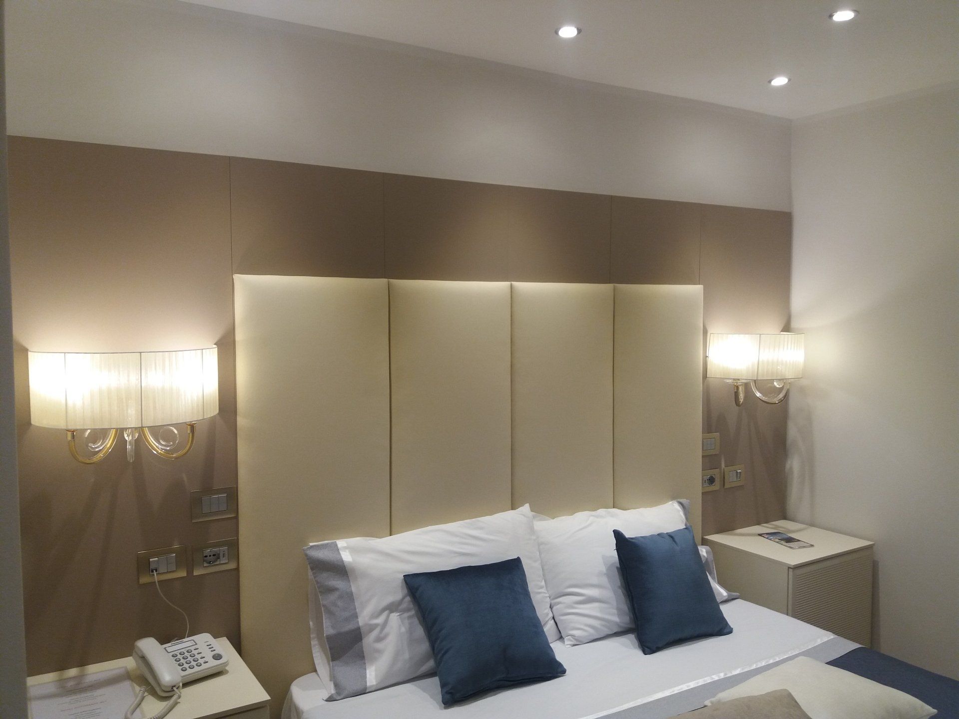 Camera da letto con un letto, due comodini, due applique e una testiera. Colori beige, bianco e blu.