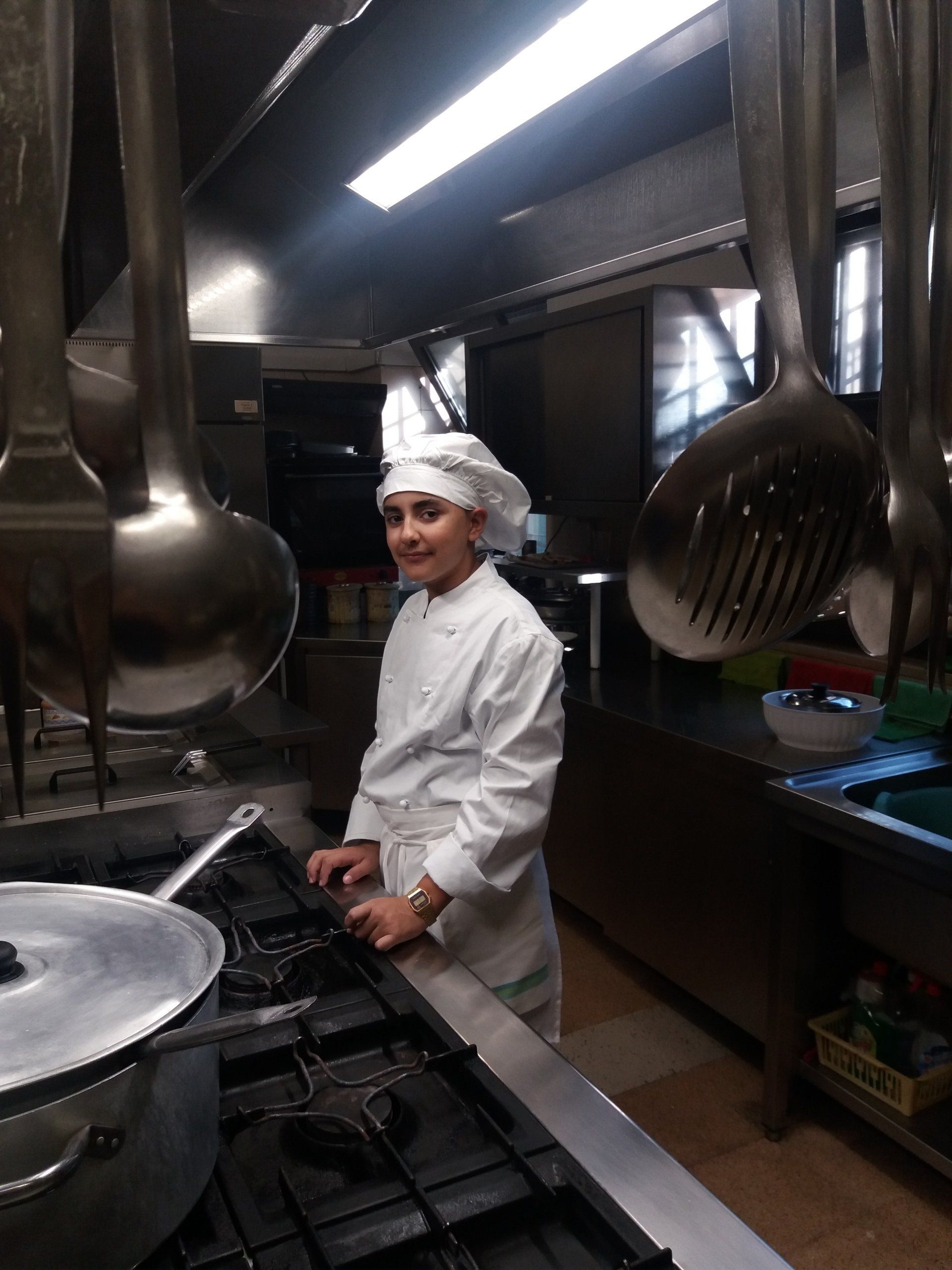 Un giovane chef in uniforme bianca e cappello è in piedi accanto ai fornelli in una cucina con utensili appesi.