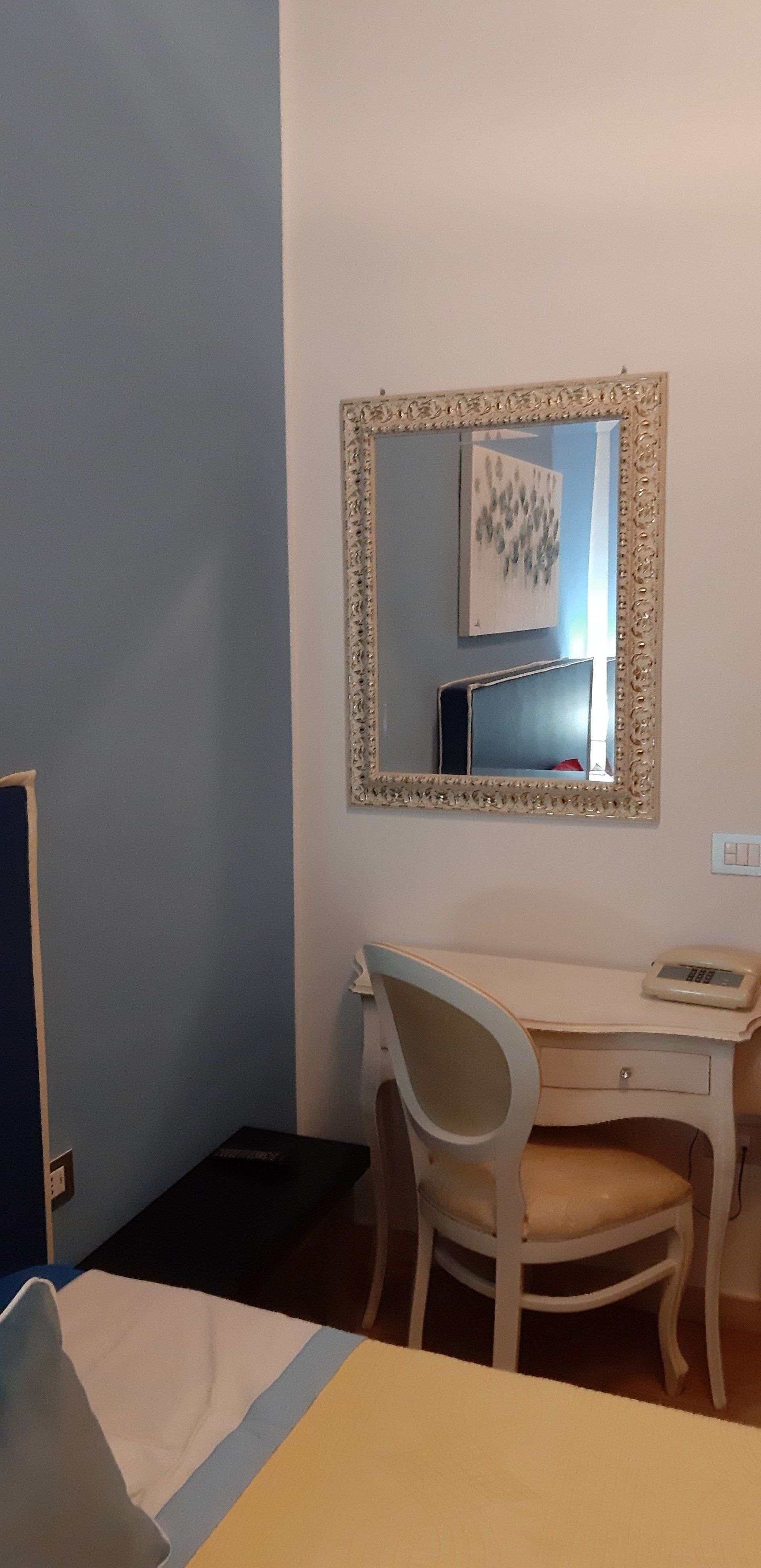 Angolo della camera da letto con scrivania, sedia, specchio e pareti blu/bianche. Il letto giallo è in primo piano.