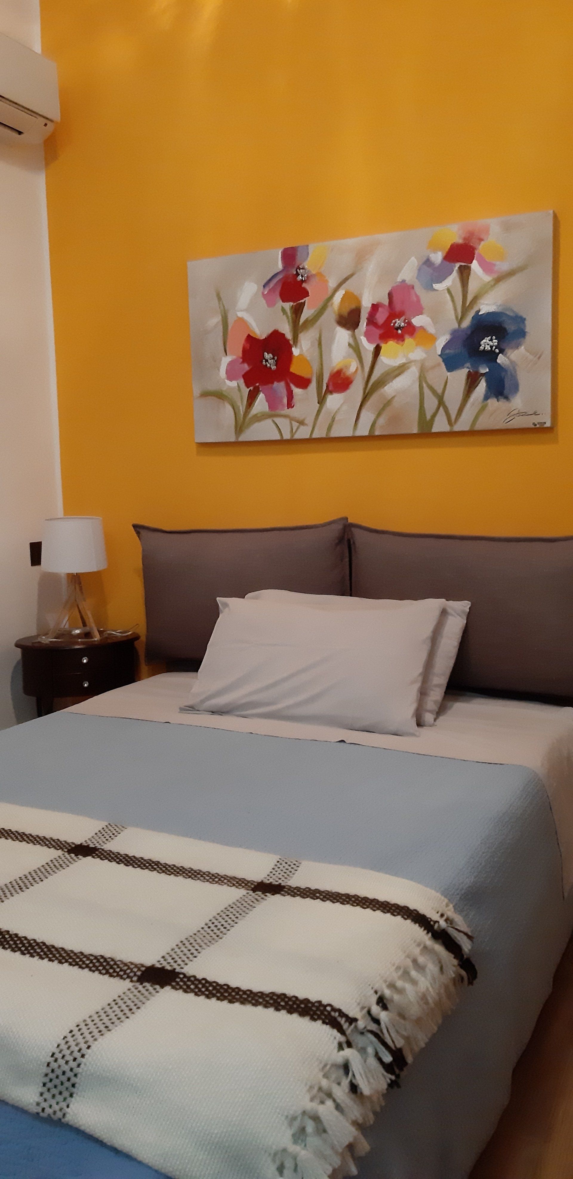 Camera da letto con parete gialla, opere d'arte floreali, letto grigio e coperta a quadri.