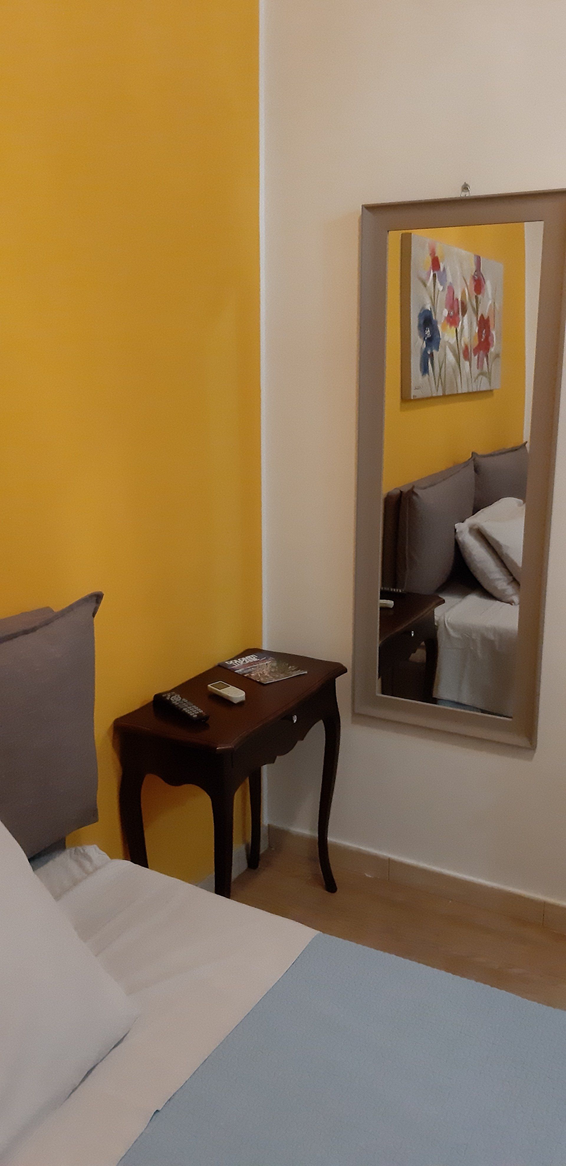 Un angolo della camera da letto con un letto, un comodino e uno specchio che riflette la stanza. Pareti gialle e bianche.