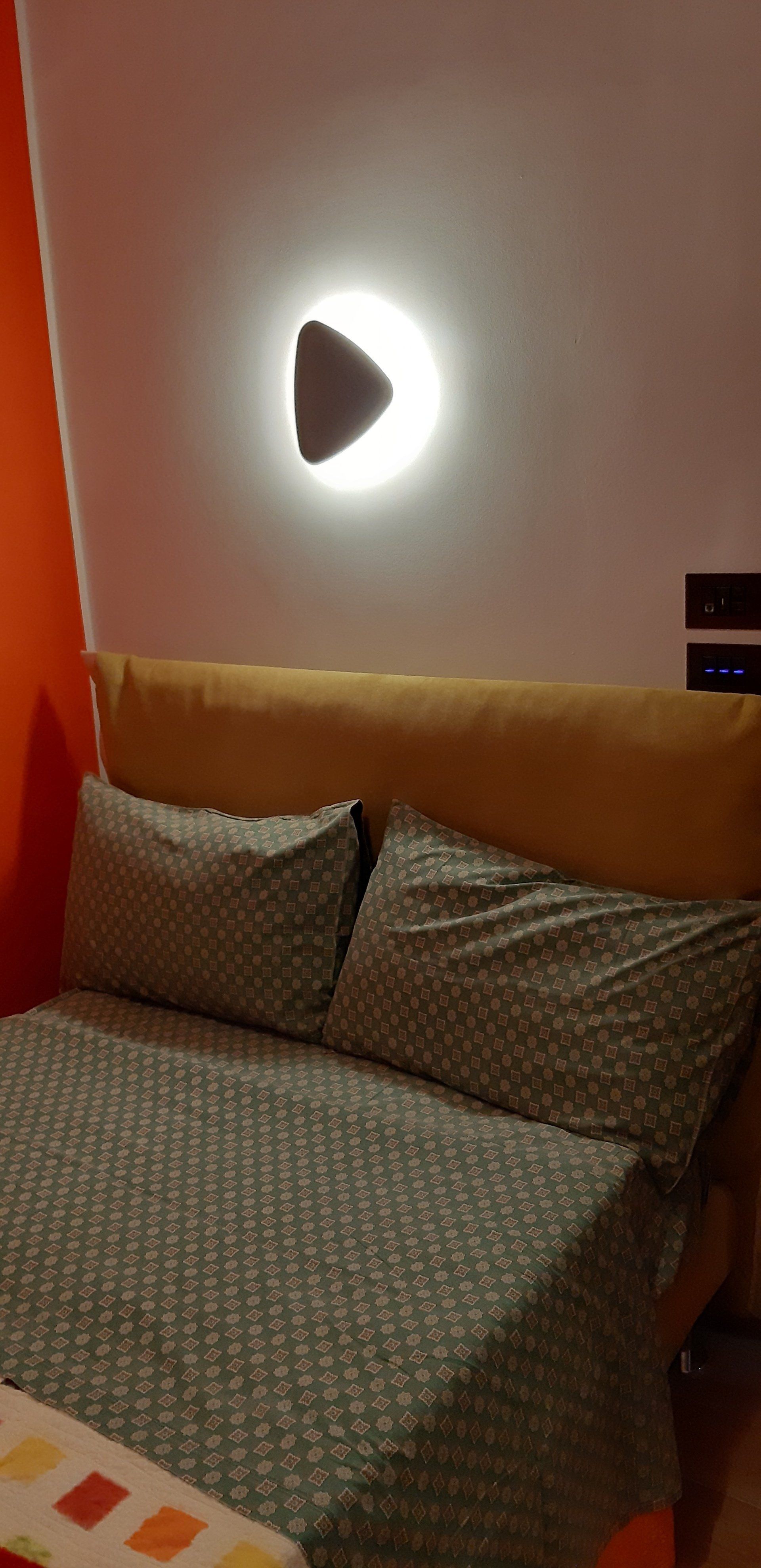 Camera da letto con letto, cuscini e una lampada da parete a forma di triangolo. La parete è bianca e arancione.