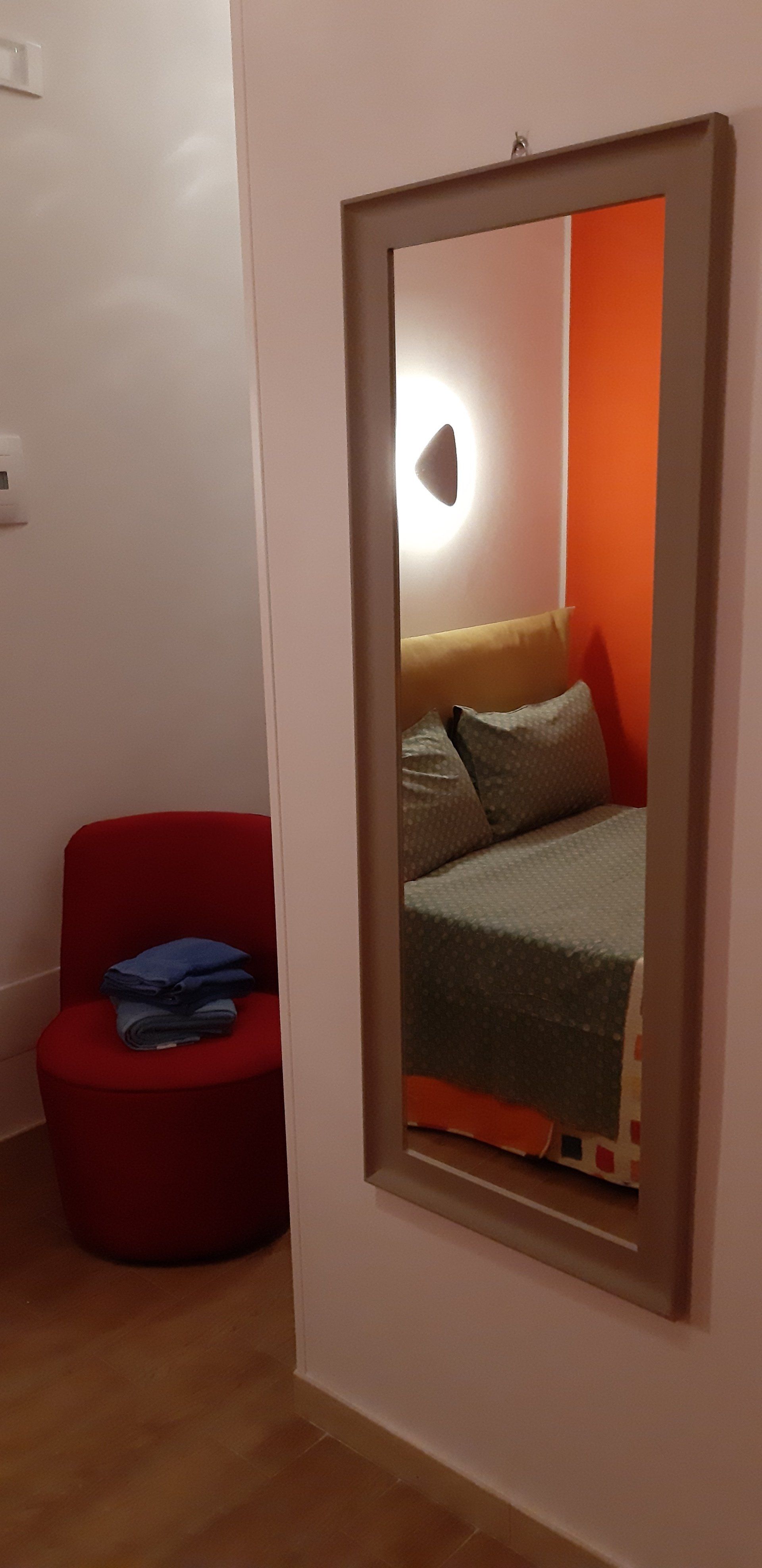 Una stanza con uno specchio che riflette un letto e una sedia rossa visibile nel corridoio.