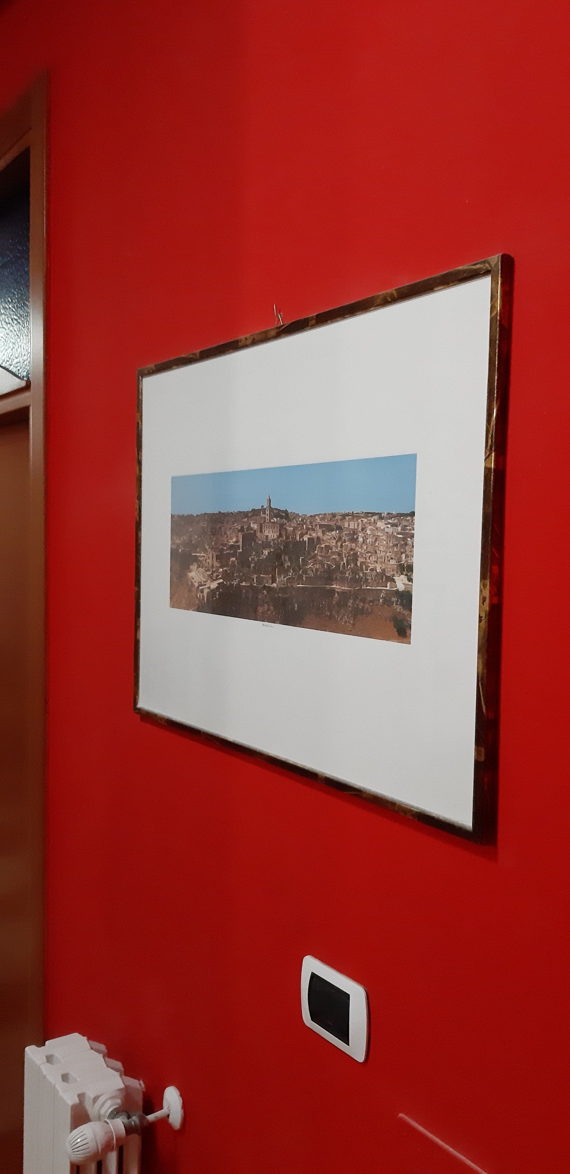 Un quadro paesaggistico incorniciato appeso a una parete rossa con un piccolo interruttore nero.
