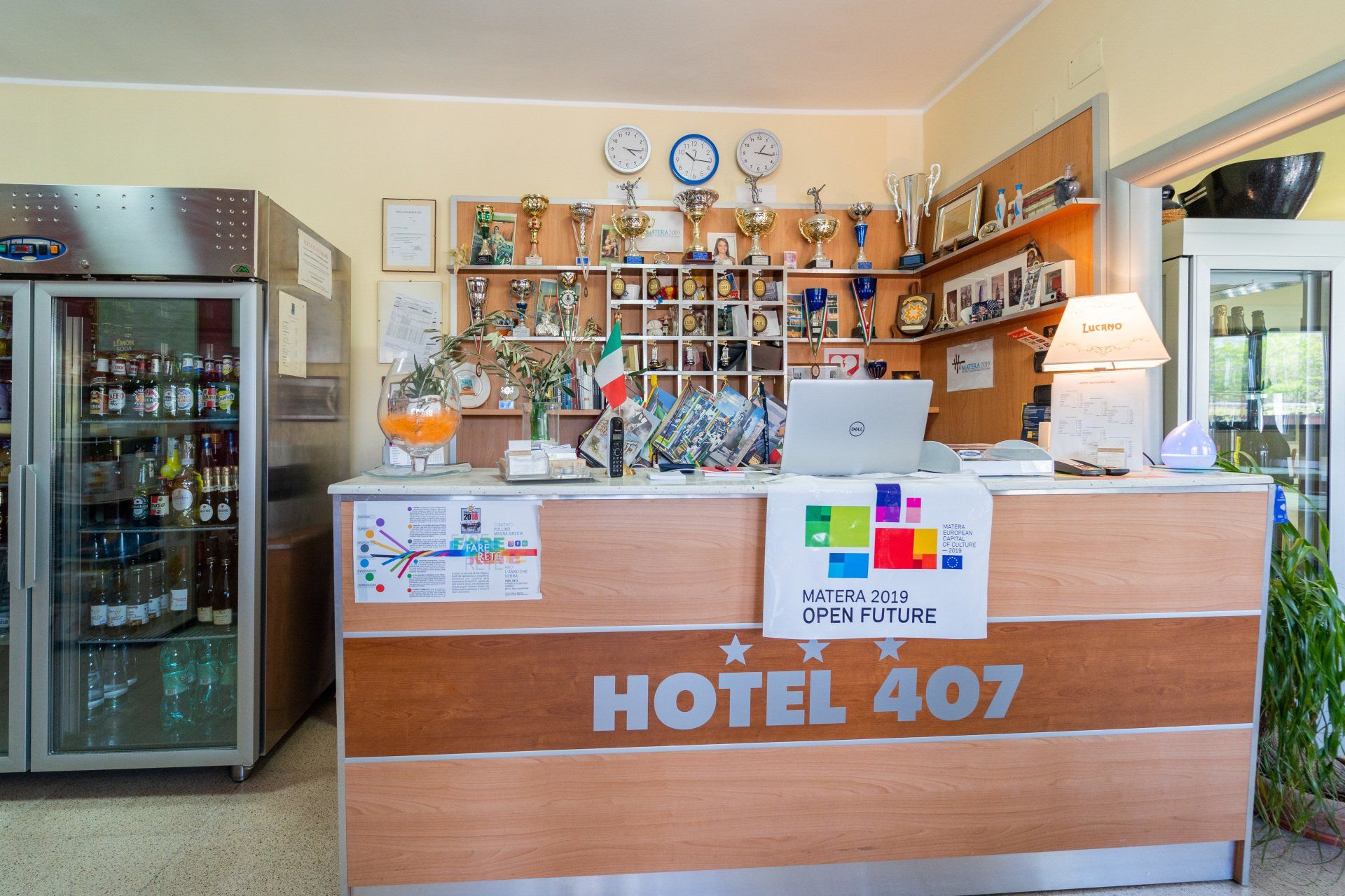 Reception dell'hotel con trofei, bandiera italiana e computer portatile. Frigoriferi portatili sulla sinistra.