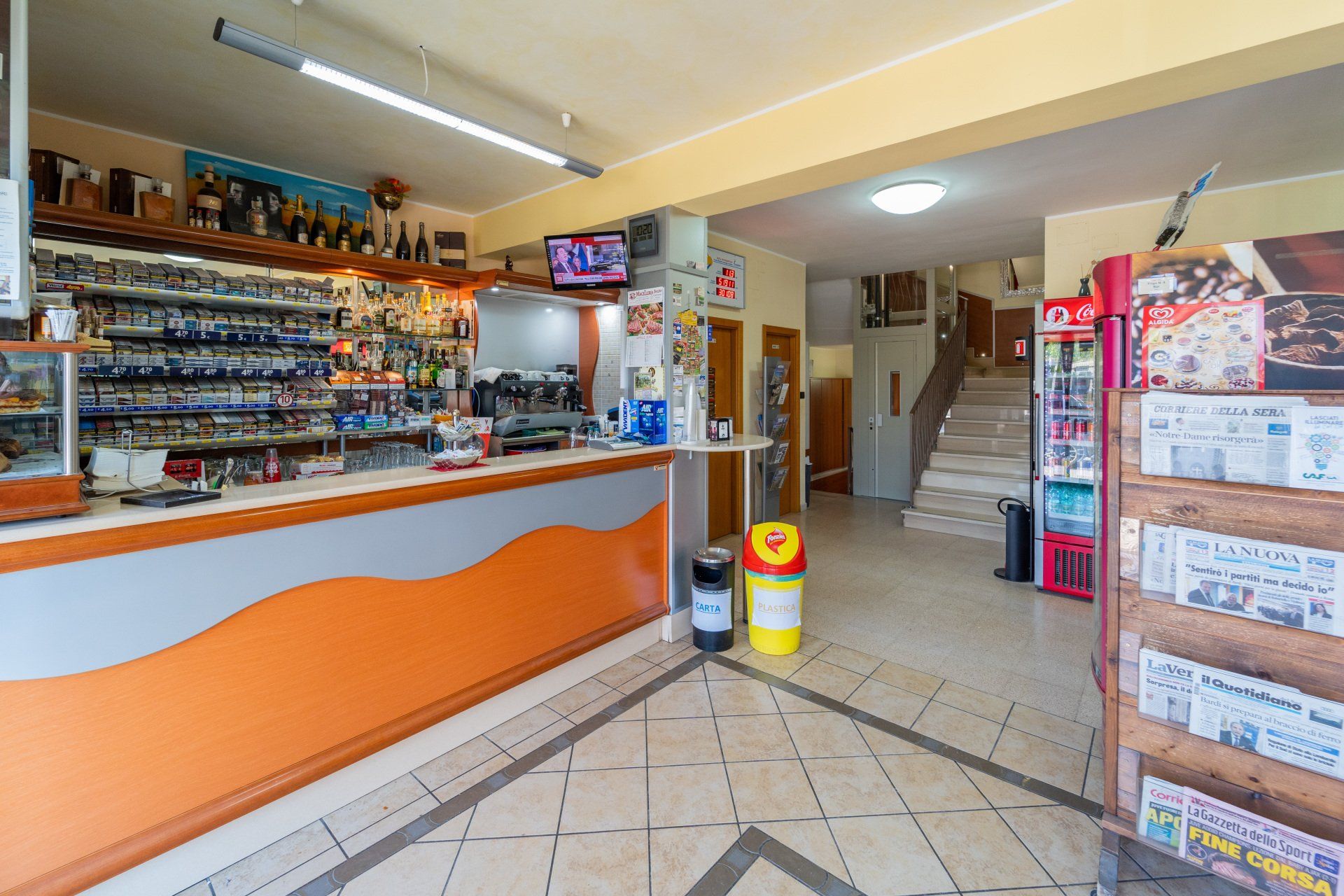 Interno di un piccolo negozio con bar, giornali e scale che portano verso l'alto; dettagli arancioni e bianchi.