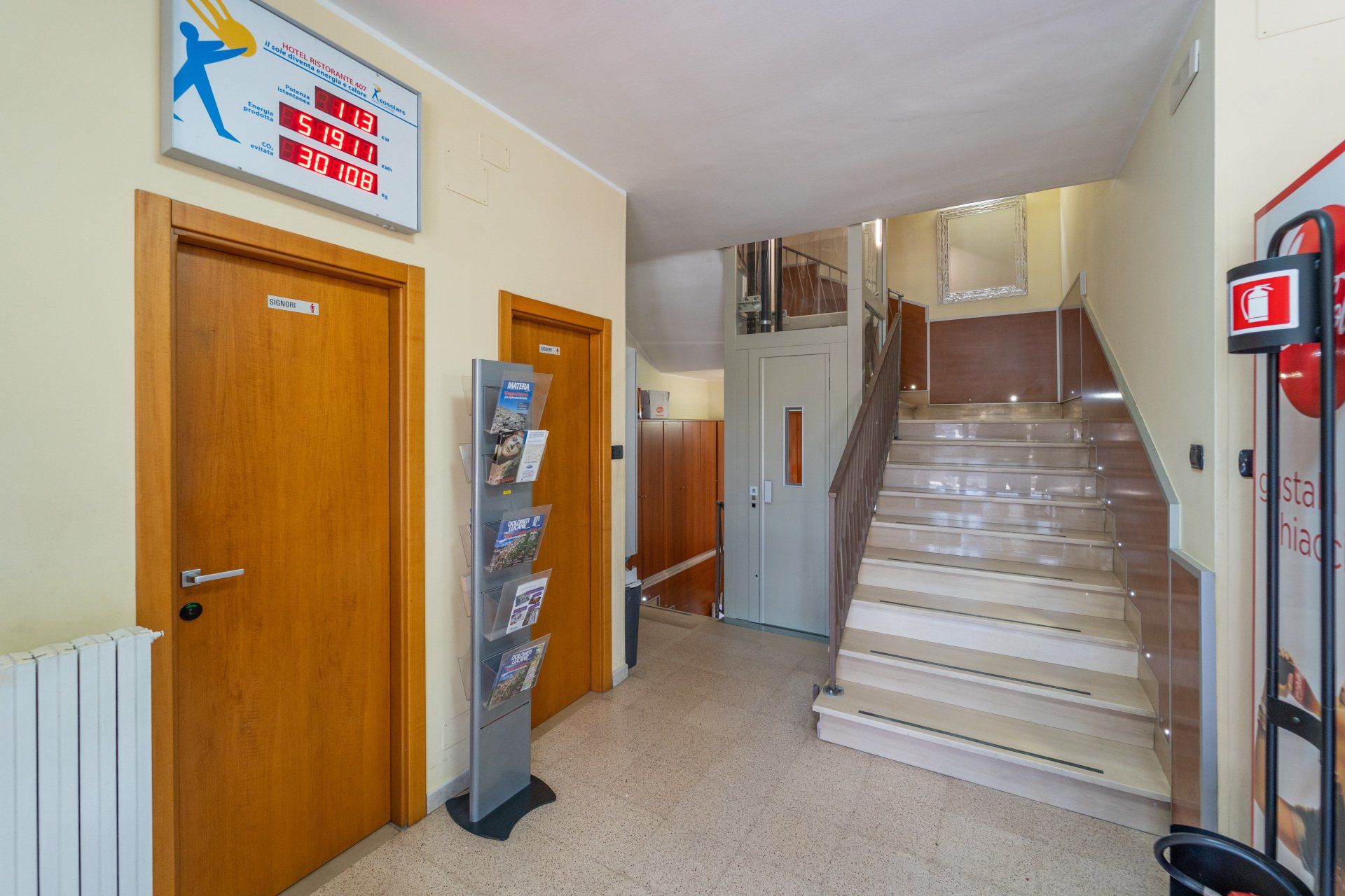 Corridoio con scale e ascensore. Due porte in legno, un porta-opuscoli e un estintore rosso. Pareti beige.