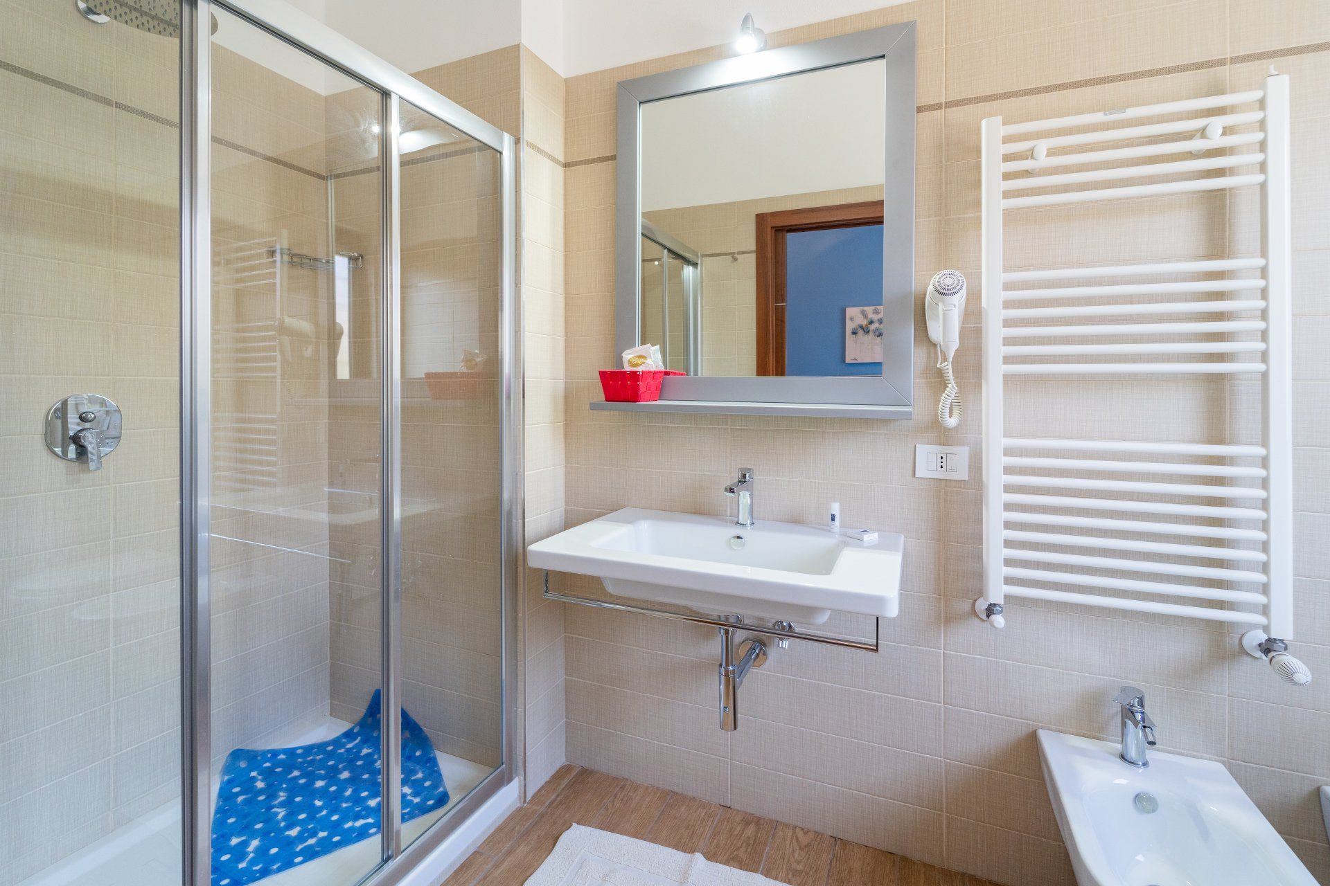 Bagno con doccia, lavandino, specchio, scaldasalviette e bidet. Pareti e pavimenti chiari.