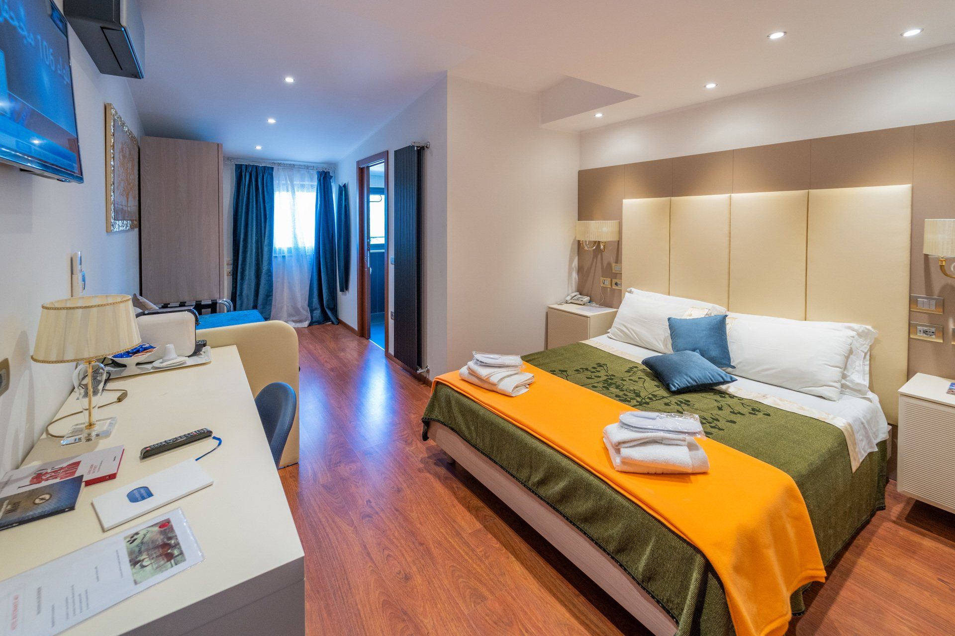 Camera d'albergo con letto, scrivania e porta-finestra che si apre su un balcone. Lenzuola verdi, arancioni e bianche. Pavimento in legno.