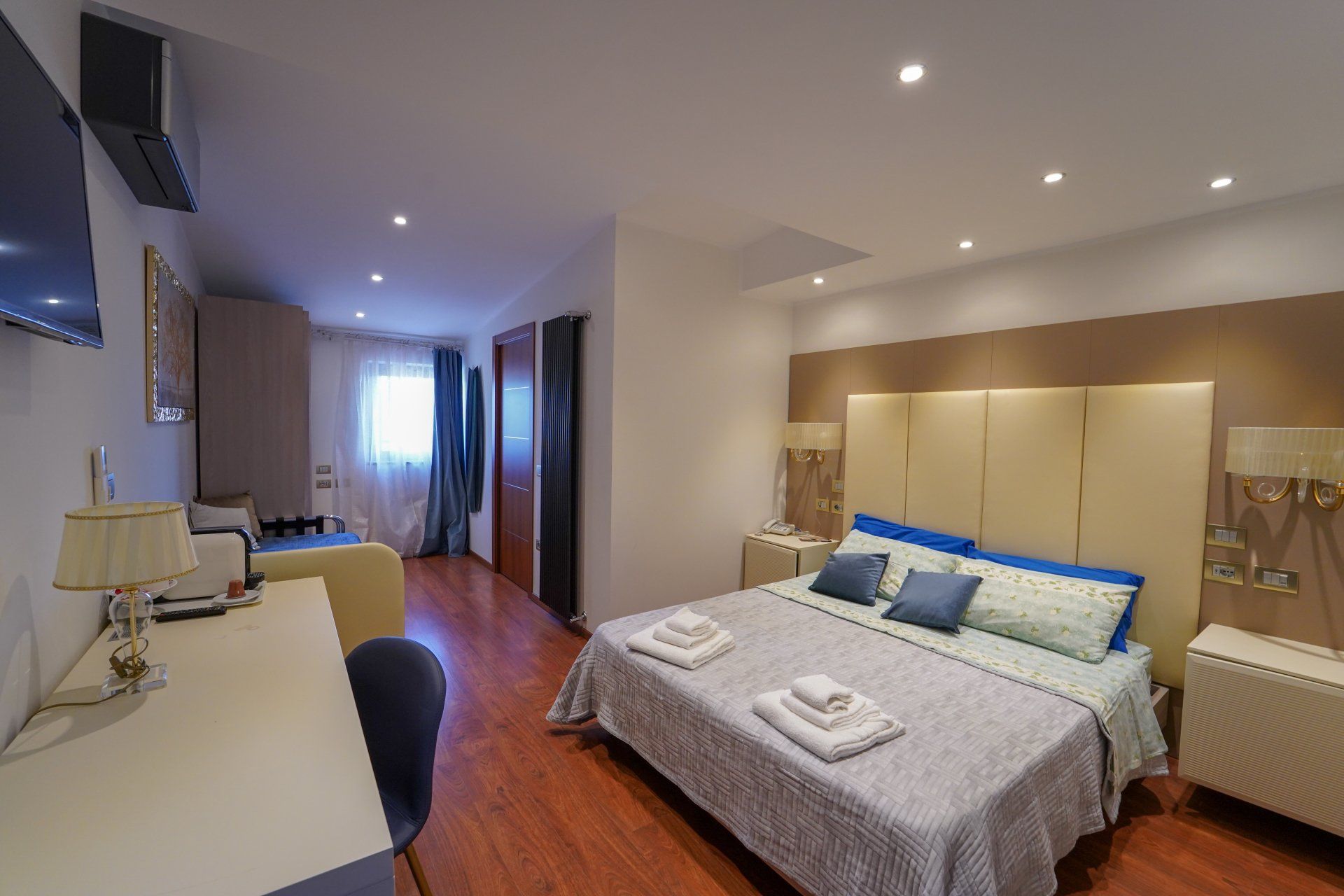 Camera da letto con letto, scrivania e zona soggiorno. Presenta pavimenti in legno, pareti neutre e un'ampia testiera.