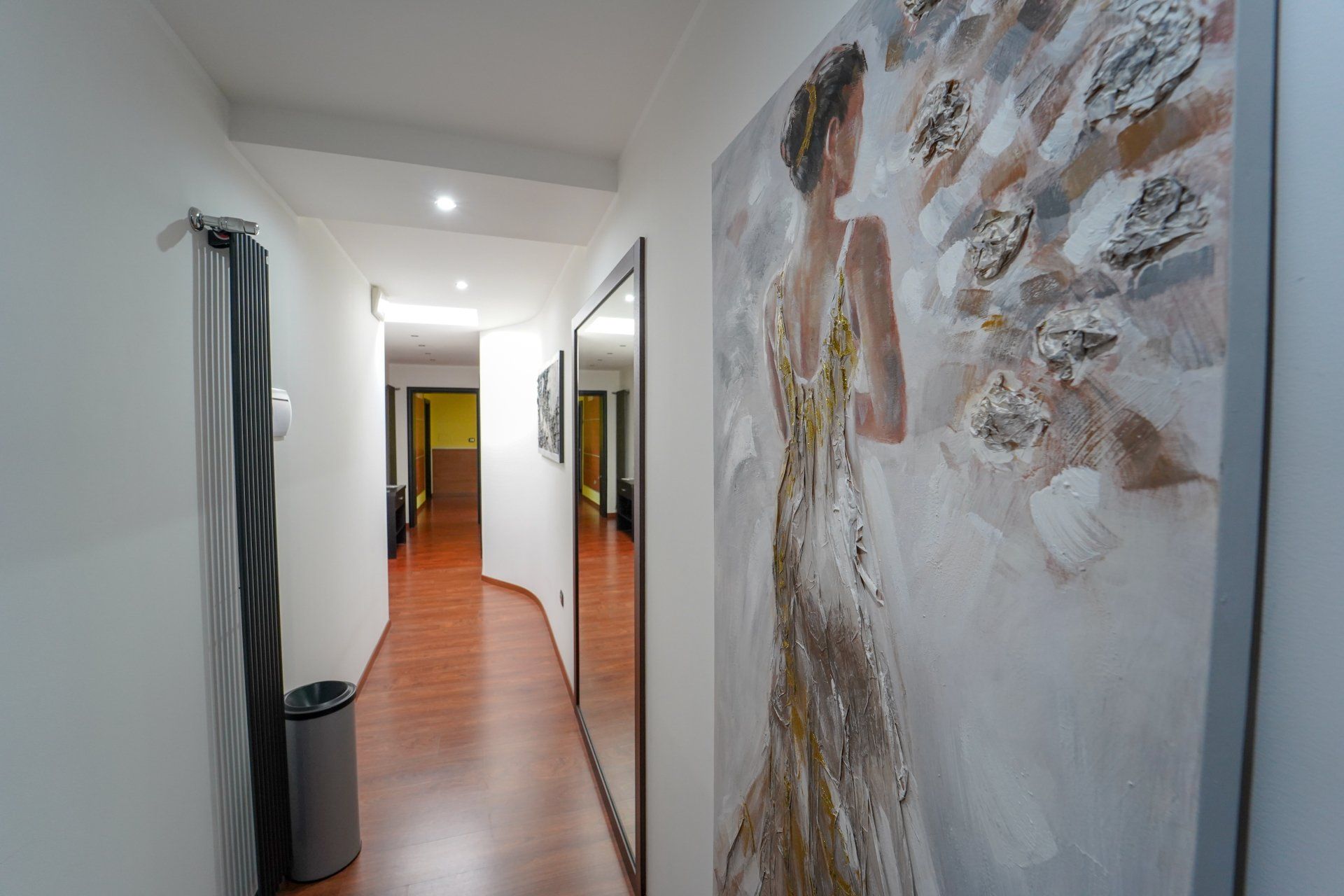 Lungo corridoio con pavimento in legno e opera d'arte raffigurante una donna in abito, parete a specchio.