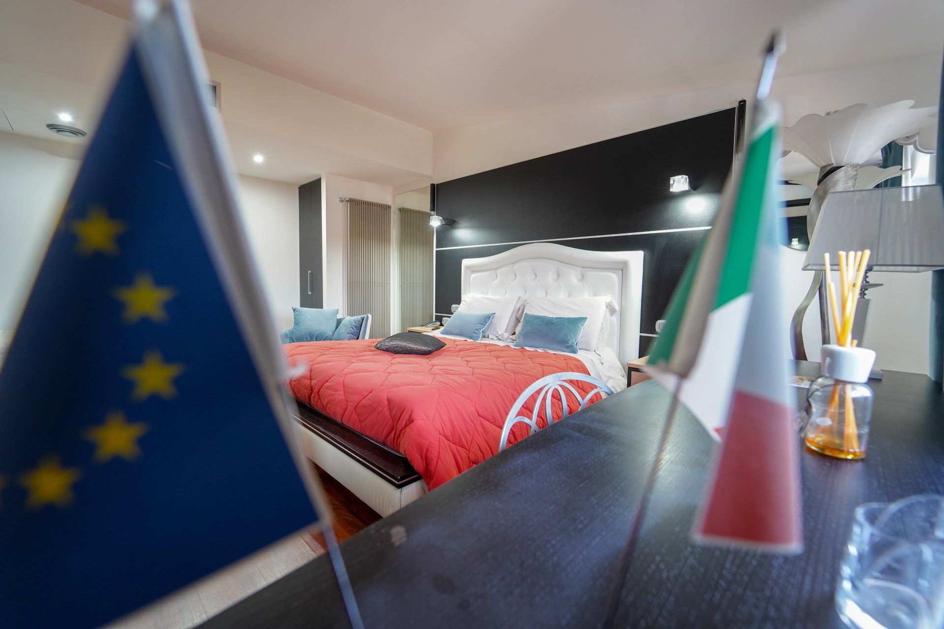 Camera da letto con bandiere italiana e dell'Unione Europea in primo piano; letto con coperta rossa.