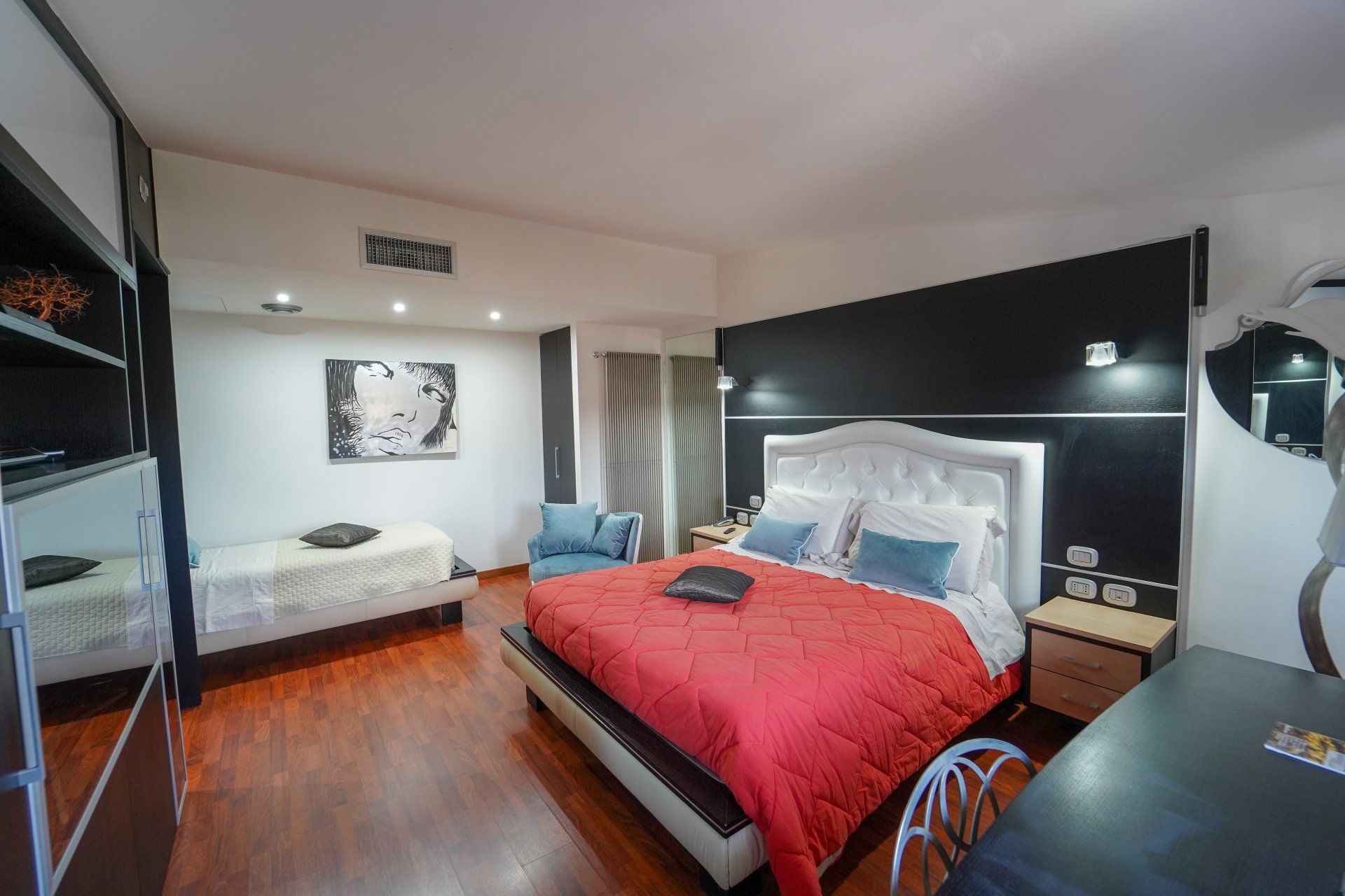 Camera da letto con copriletto rosso, letto singolo, poltrona, scrivania e parete con accenti scuri.