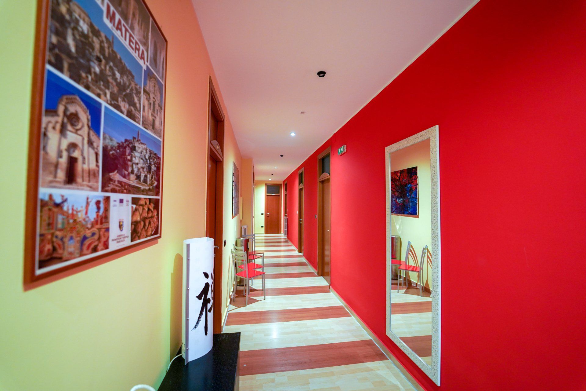 Lungo corridoio con pareti gialle e rosse, pavimento a strisce, specchio, opere d'arte.
