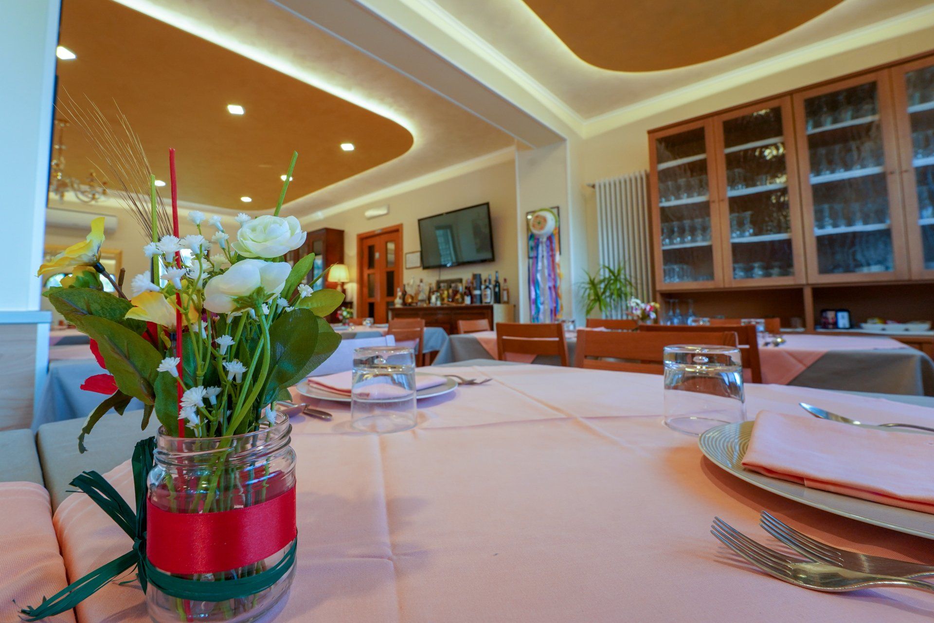 Tavolo da ristorante apparecchiato per cenare, con centrotavola floreale, tovaglia rosa pastello e mobili in legno.