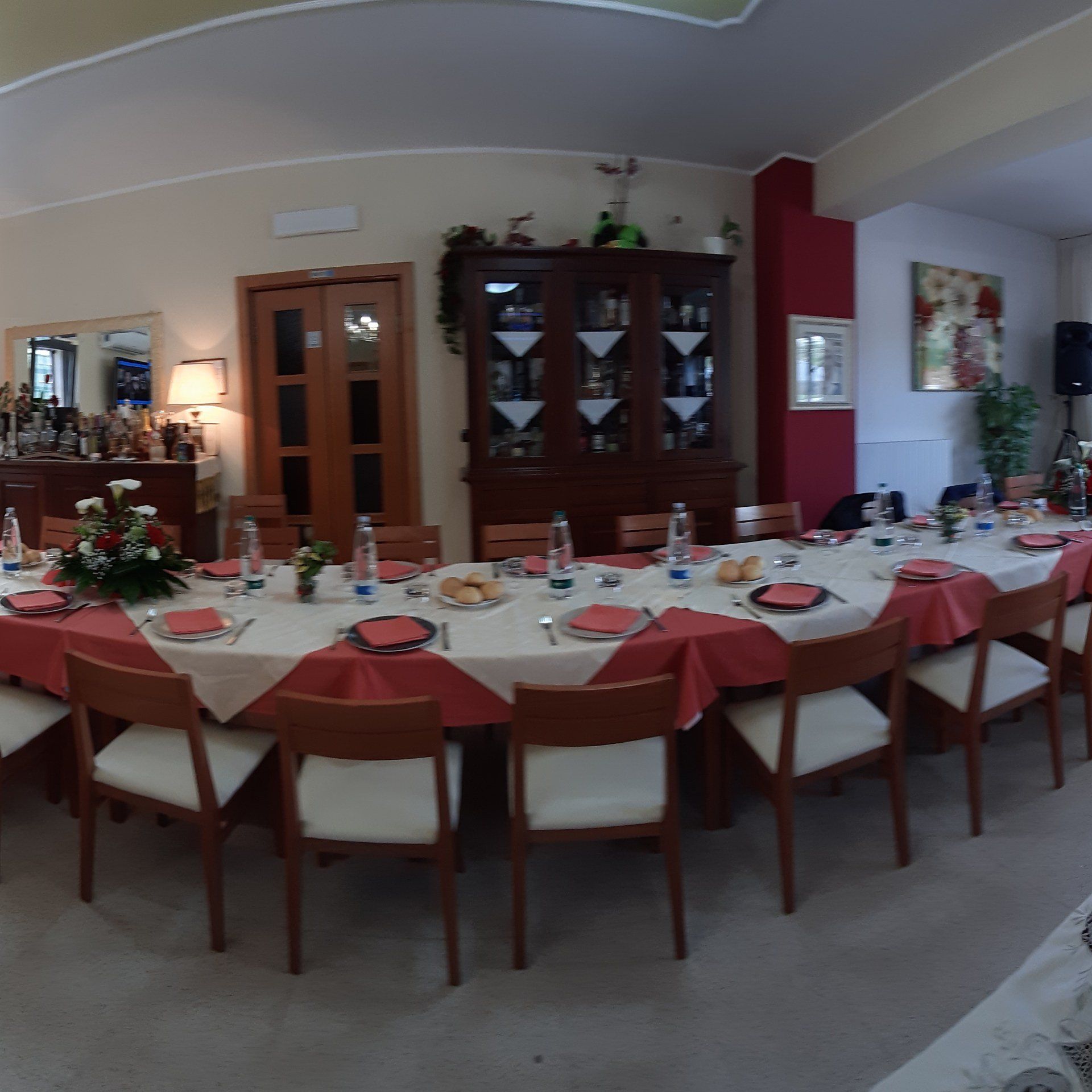 Lungo tavolo apparecchiato per un pasto in una sala da pranzo con dettagli rossi, tovaglia bianca e sedie in legno.