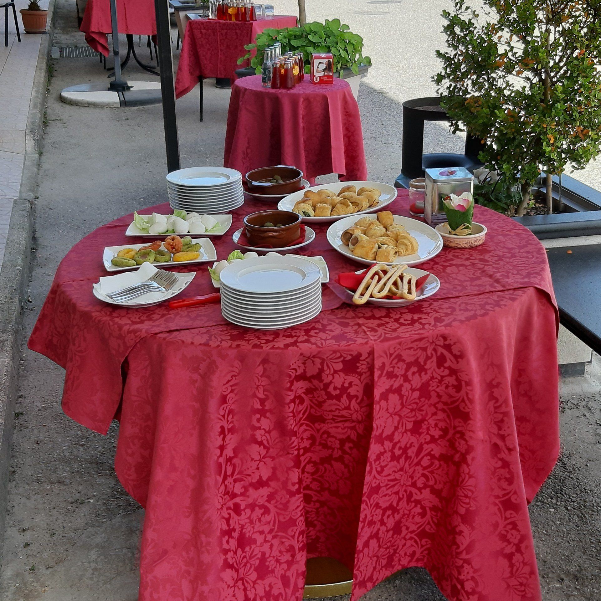 Tavoli all'aperto apparecchiati per un pasto, tovaglie rosse, vari piatti, piatti e bicchieri.