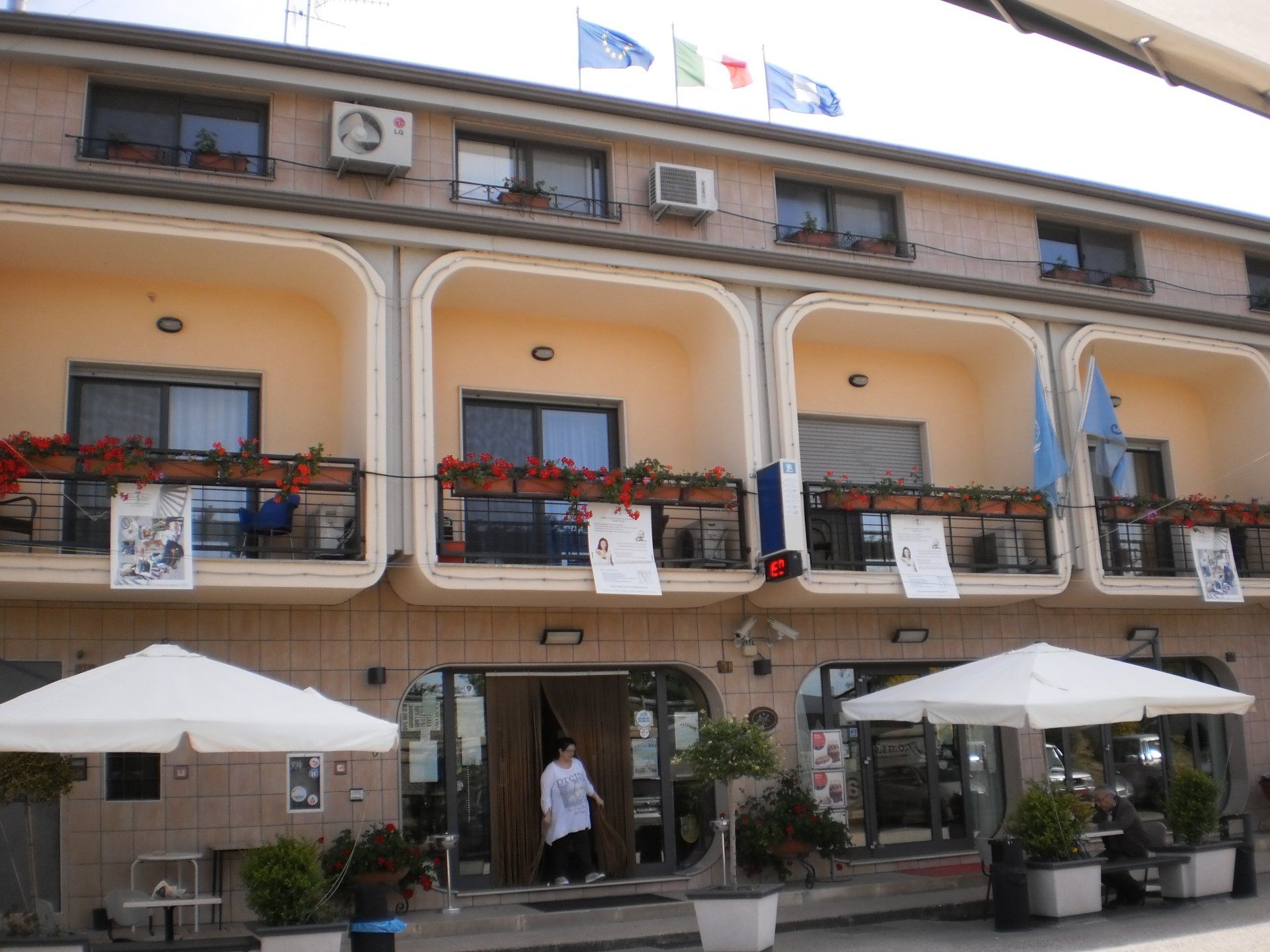 Esterno dell'hotel con balconi ad arco, bandiere, tende da sole e una persona all'ingresso.