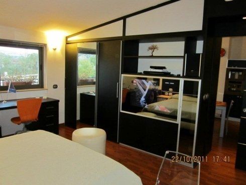 Camera da letto con armadio nero, specchio, scrivania e letto bianco. Interni luminosi e moderni con finestra.