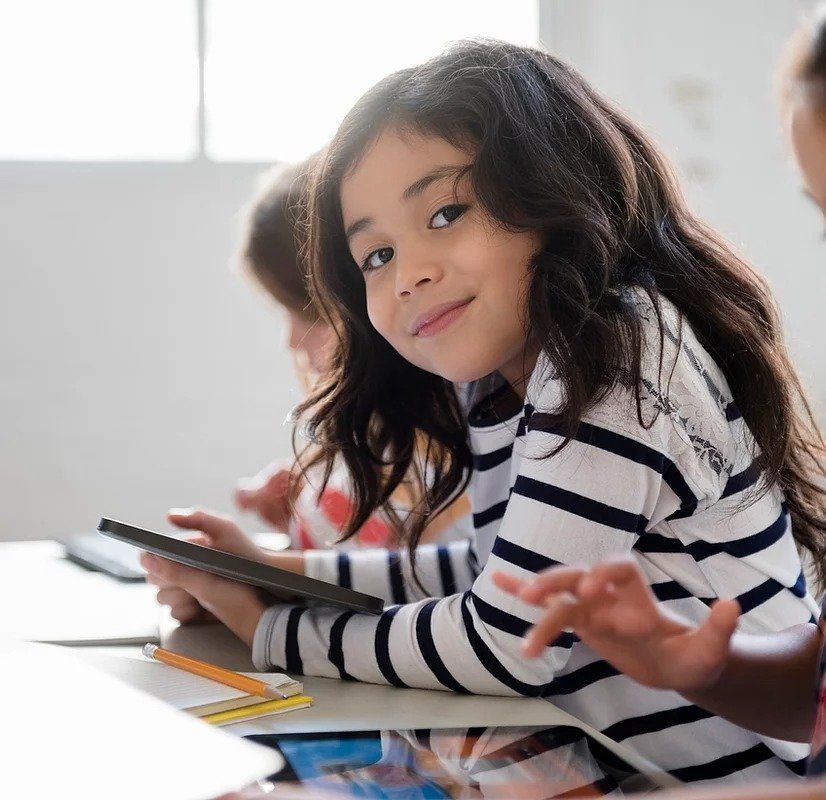 A little girl in a striped shirt is sitting at a desk holding a tablet