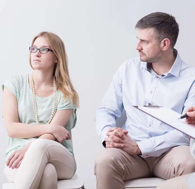 A man and a woman are sitting in chairs while a man holds a clipboard.