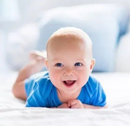 A baby is laying on his stomach on a bed and smiling.