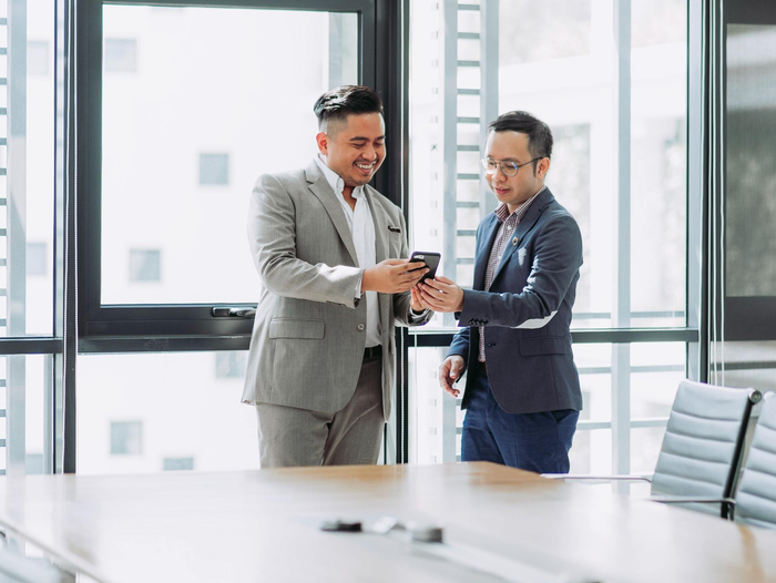 Two men in suits are standing next to each other in a conference room looking at a cell phone.